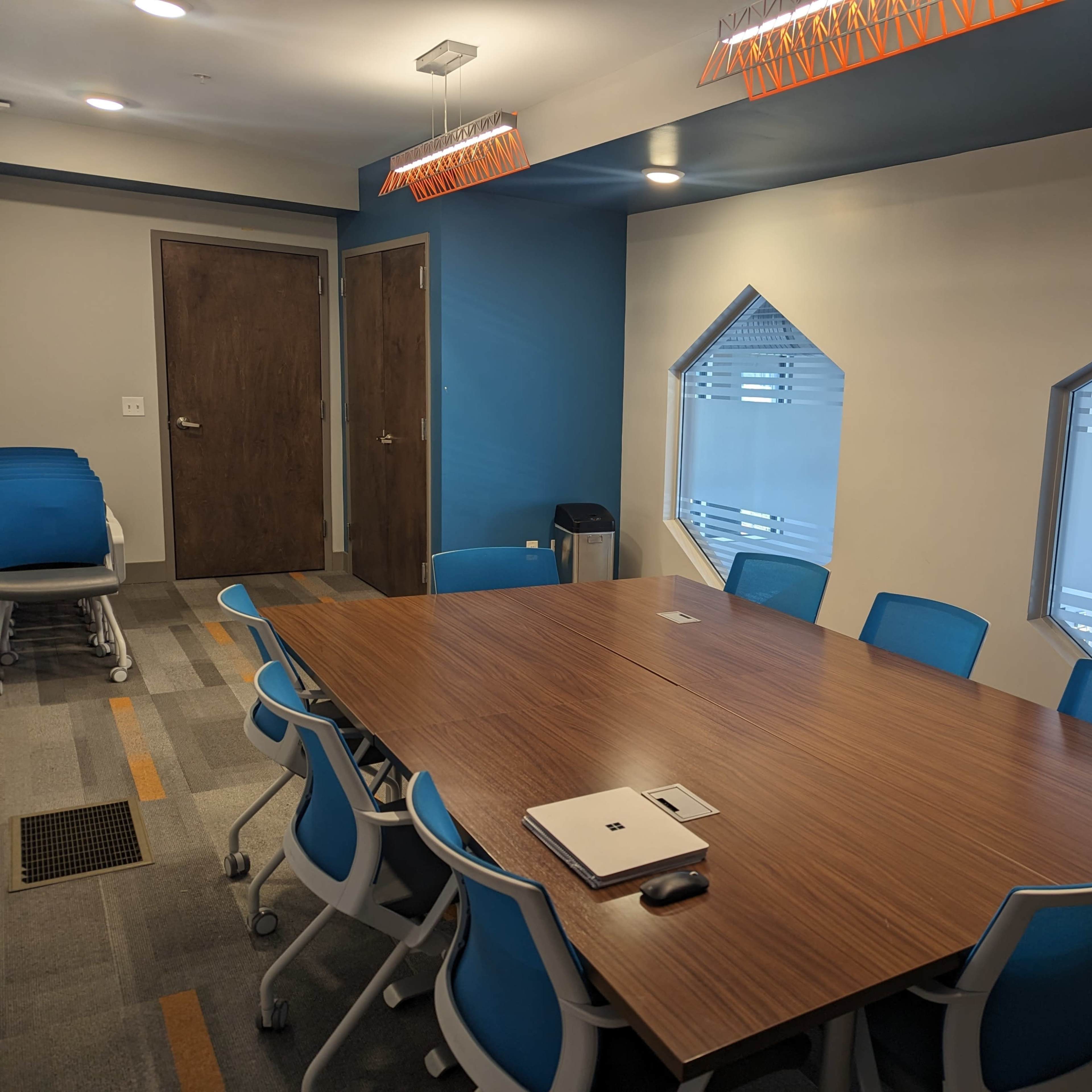 A conference room features a large wooden table surrounded by blue chairs, with two unique windows and overhead lighting fixtures.