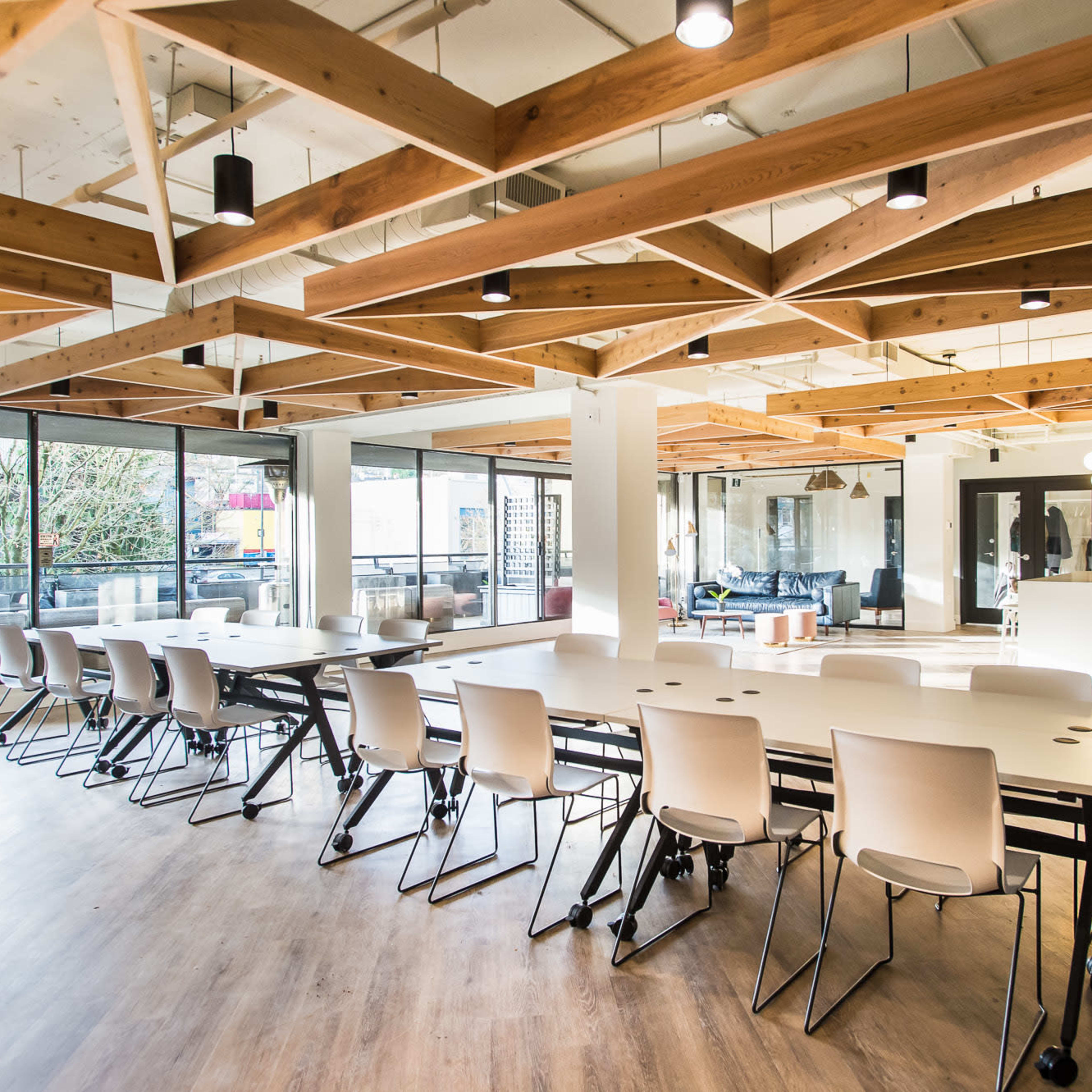 A spacious modern office with a large table, multiple chairs, and wooden beams in the ceiling, illuminated by natural light from large windows.