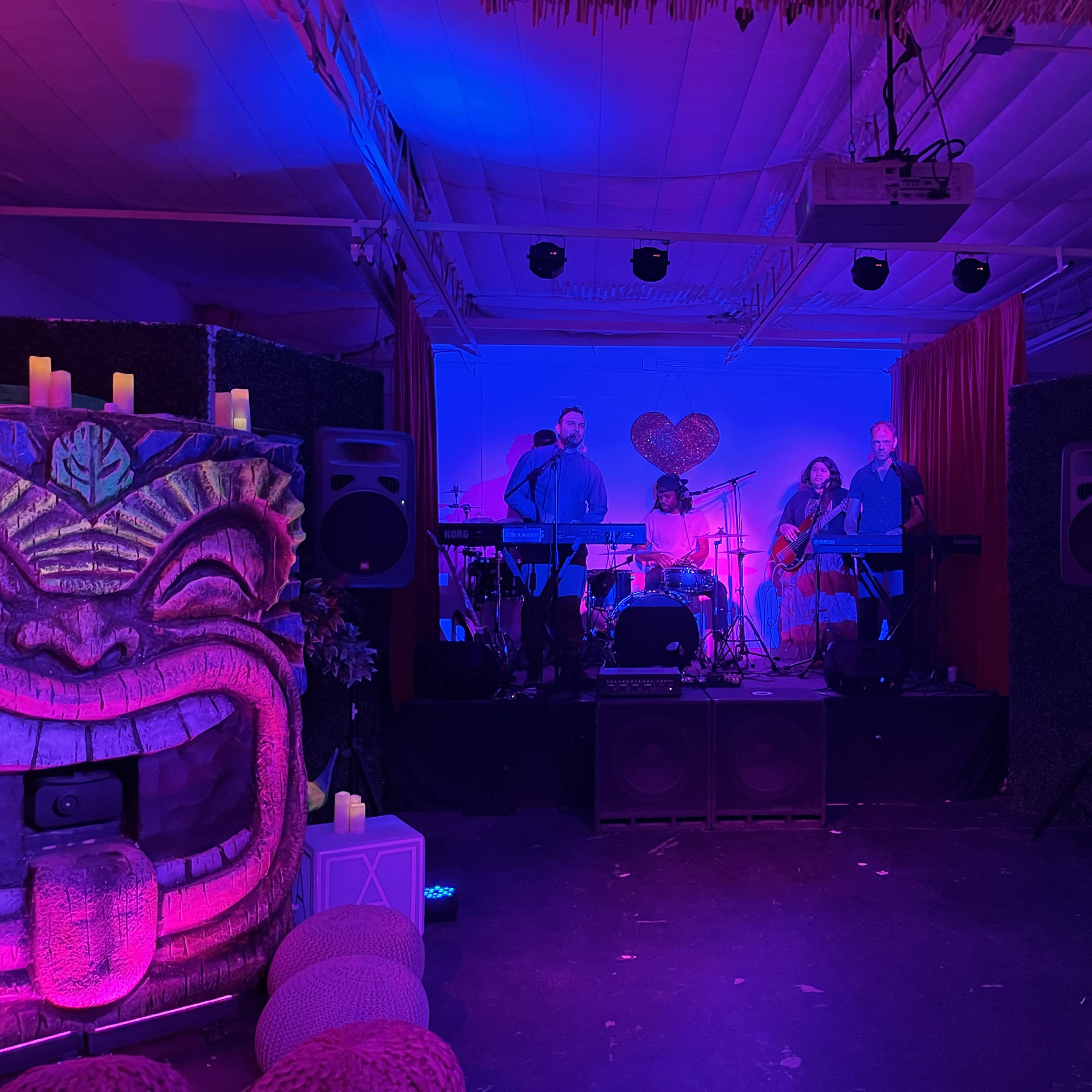 A band performs on a small stage decorated with tropical-themed artwork and ambient lighting.