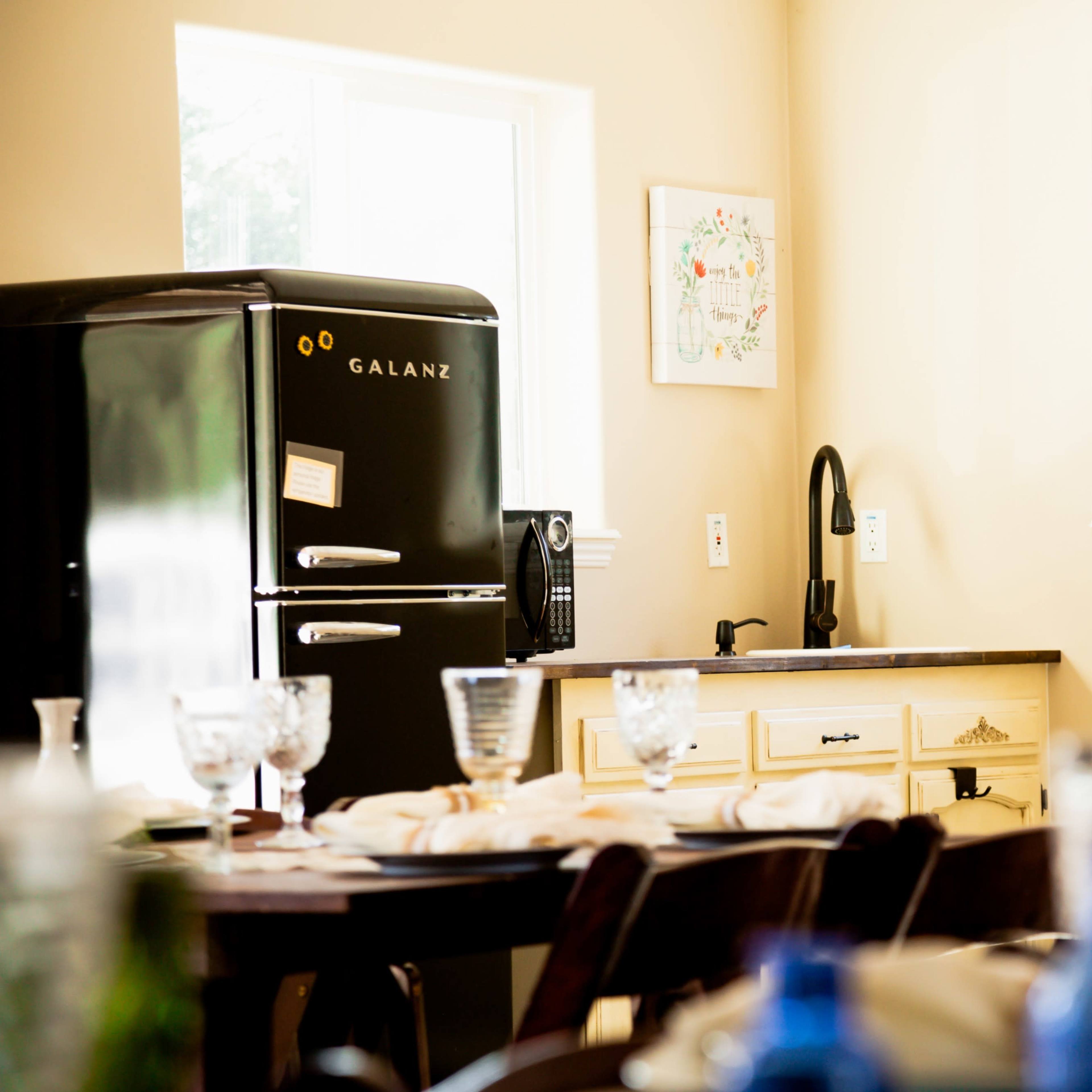 A black refrigerator stands beside a sink in a kitchen with light-colored walls and a wooden dining table set with glassware.