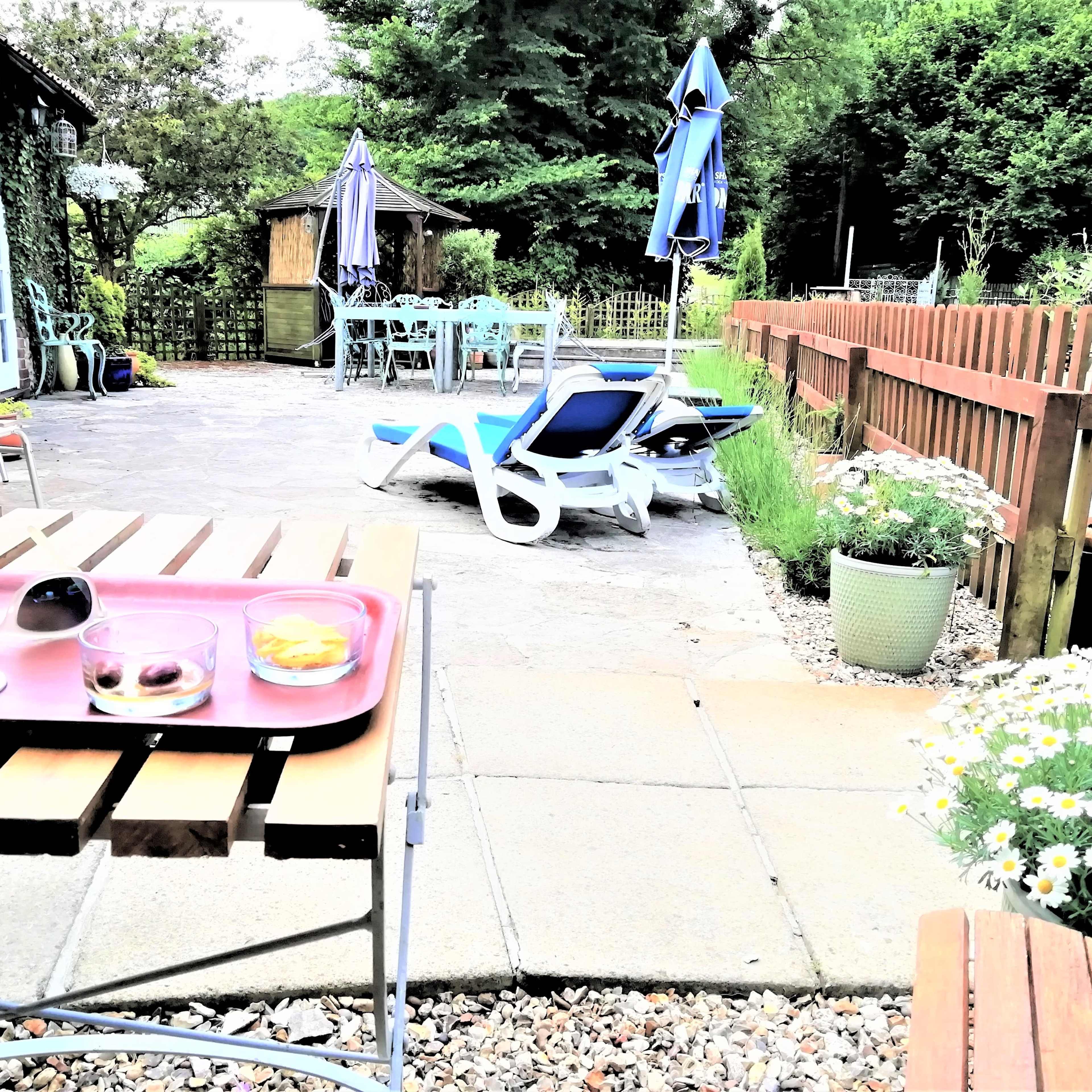 A garden patio with a table set for refreshments, lounge chairs, and a shaded area surrounded by greenery.