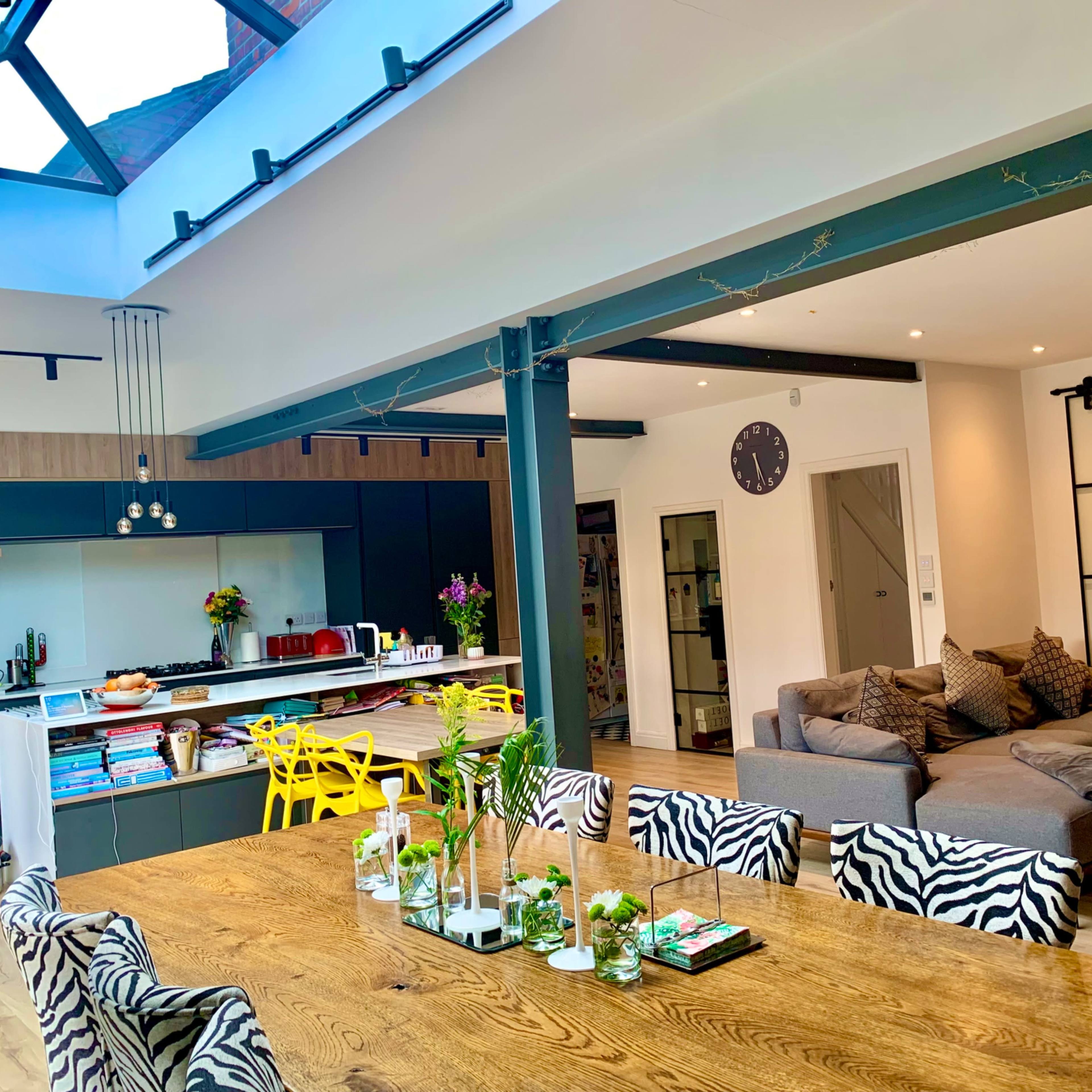 A modern kitchen and dining area with a large wooden table, vibrant yellow chairs, and a glass roof that allows natural light to enter the space.