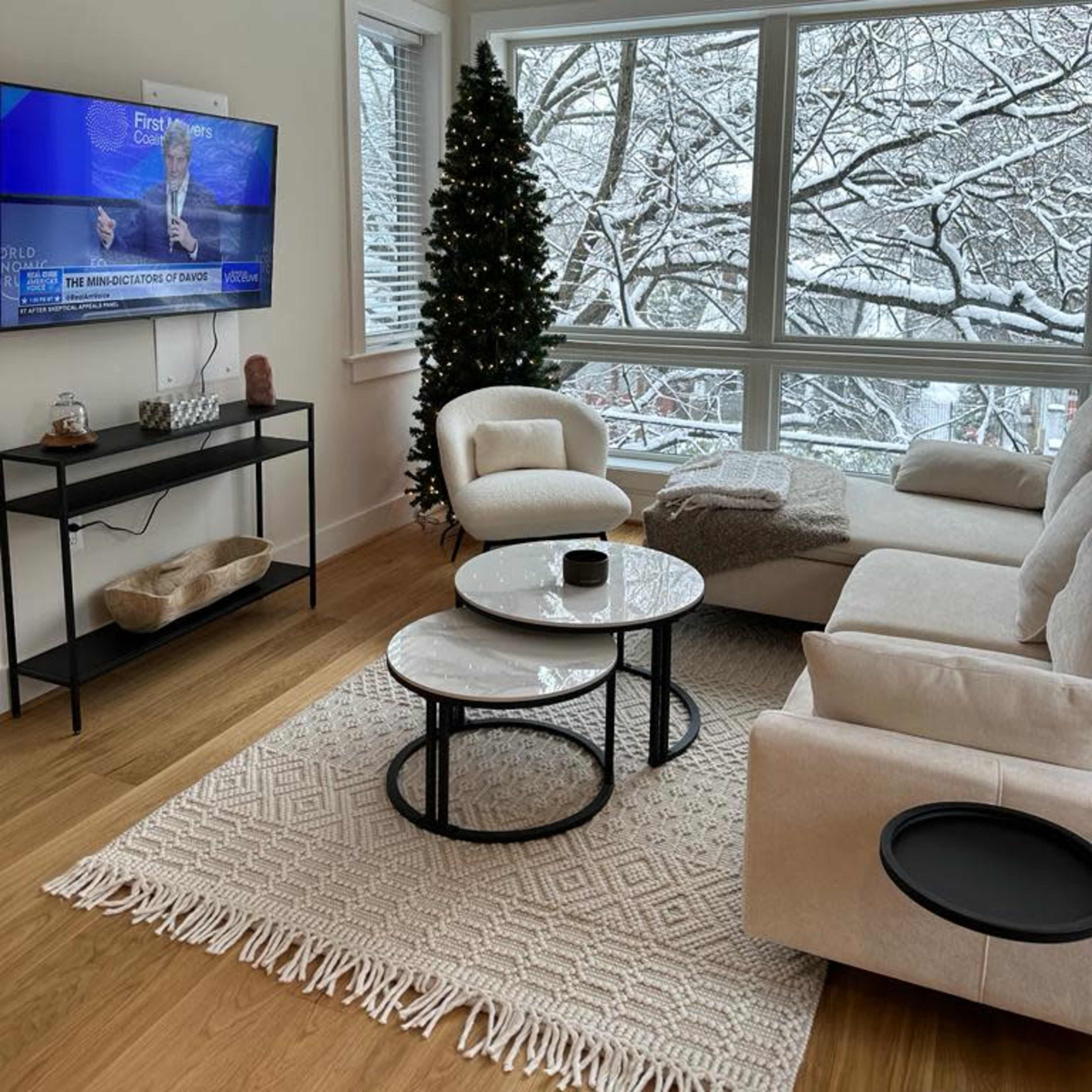 A cozy living room features a light-colored sofa, a round coffee table, and a small Christmas tree beside a large window overlooking a snowy landscape.