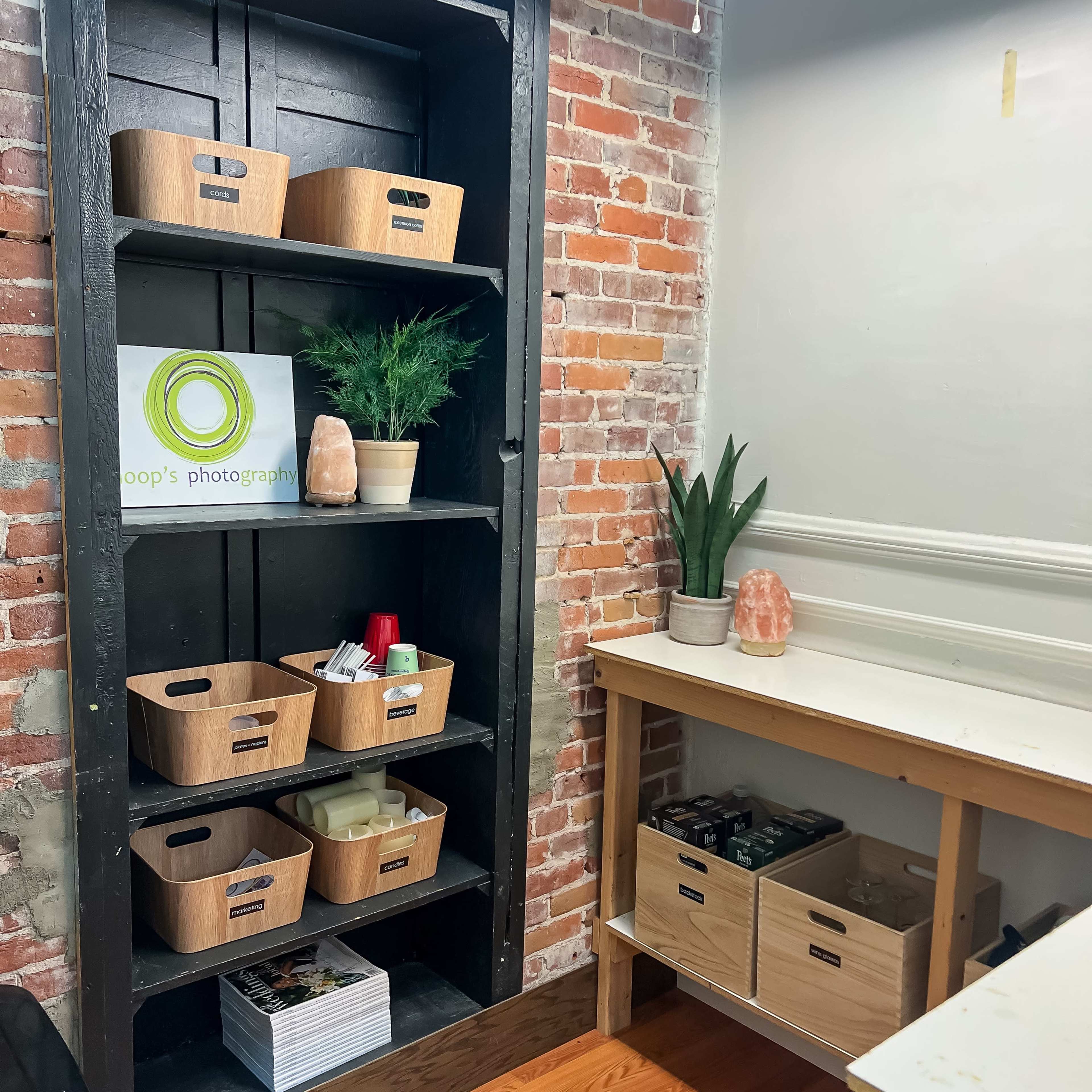 A shelving unit filled with woven baskets and plants, alongside a wooden countertop and storage boxes in a room with exposed brick walls.
