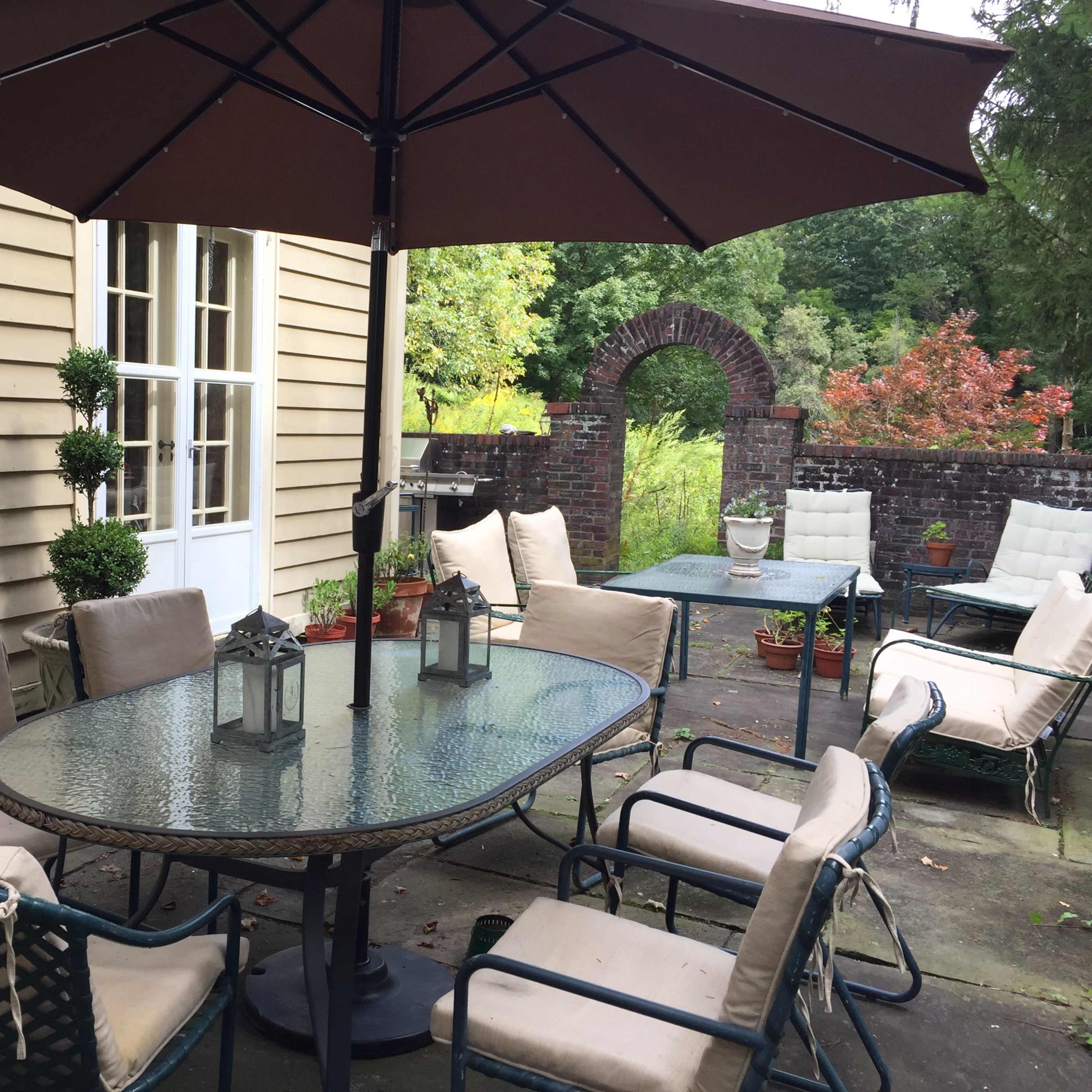A patio area features a glass table surrounded by metal chairs, with an umbrella overhead and potted plants nearby.