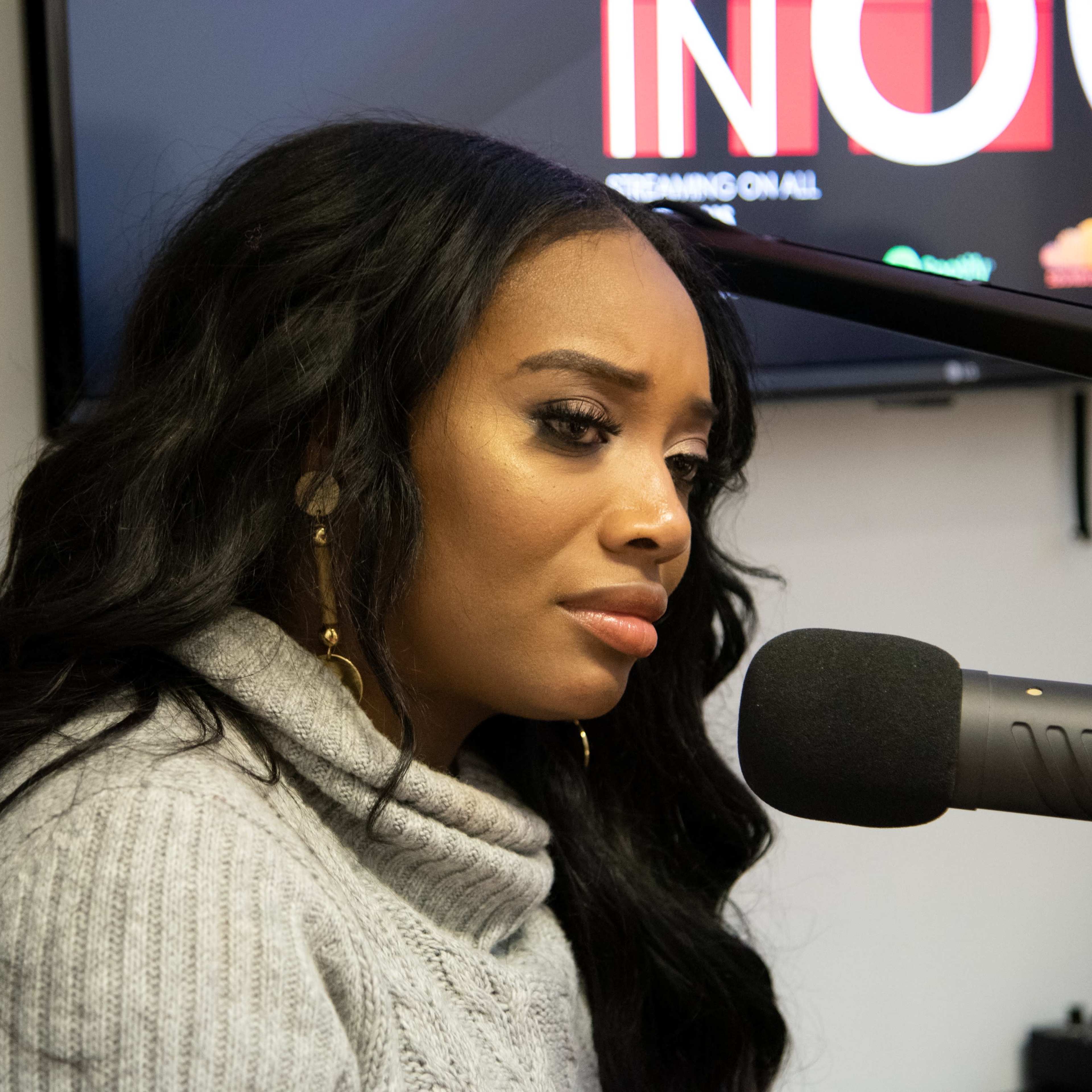 A woman with long, wavy hair sits in front of a microphone, appearing deep in thought during a recording session.