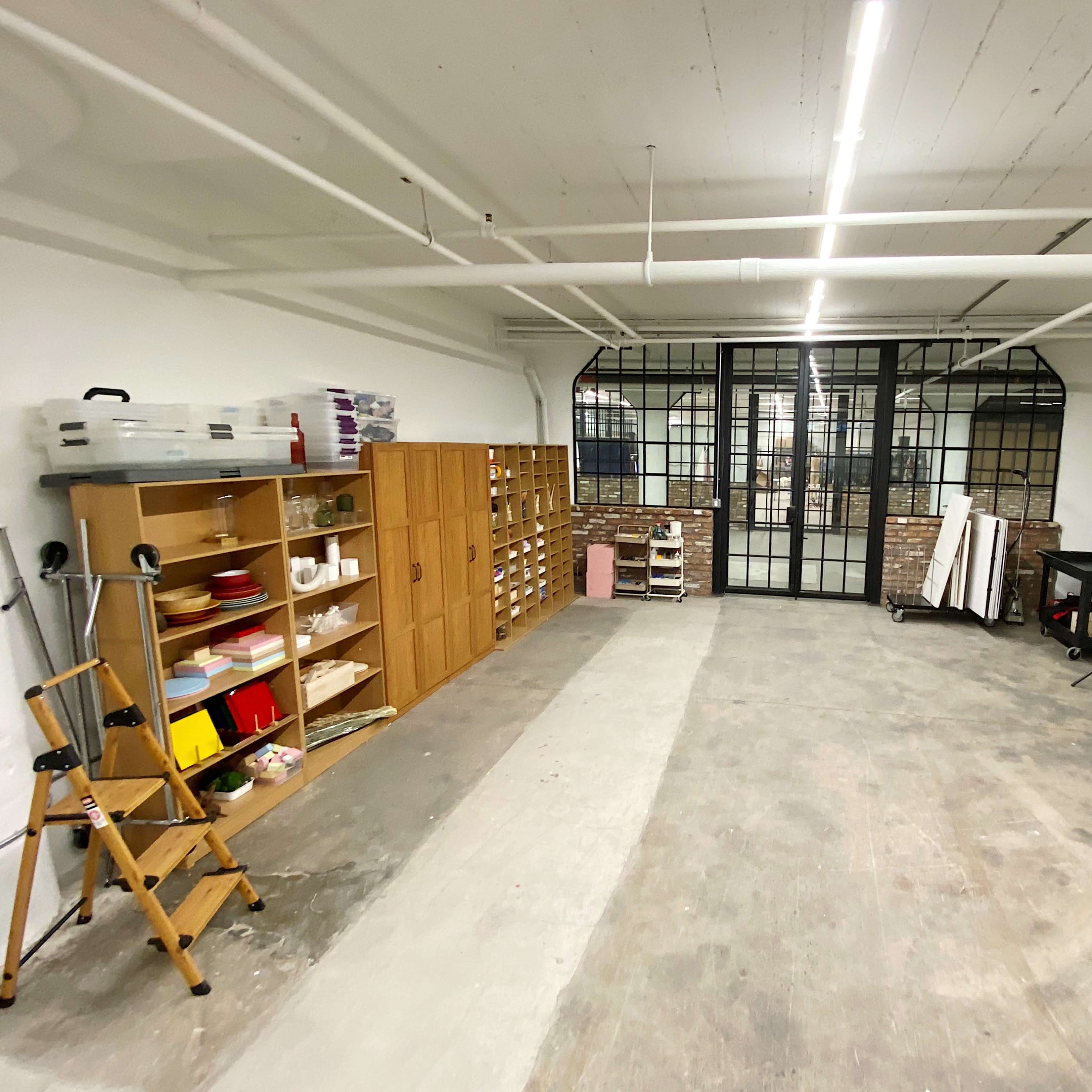 A spacious, empty room with a wooden shelving unit on the left, a table on the right, and large glass windows at the far end.