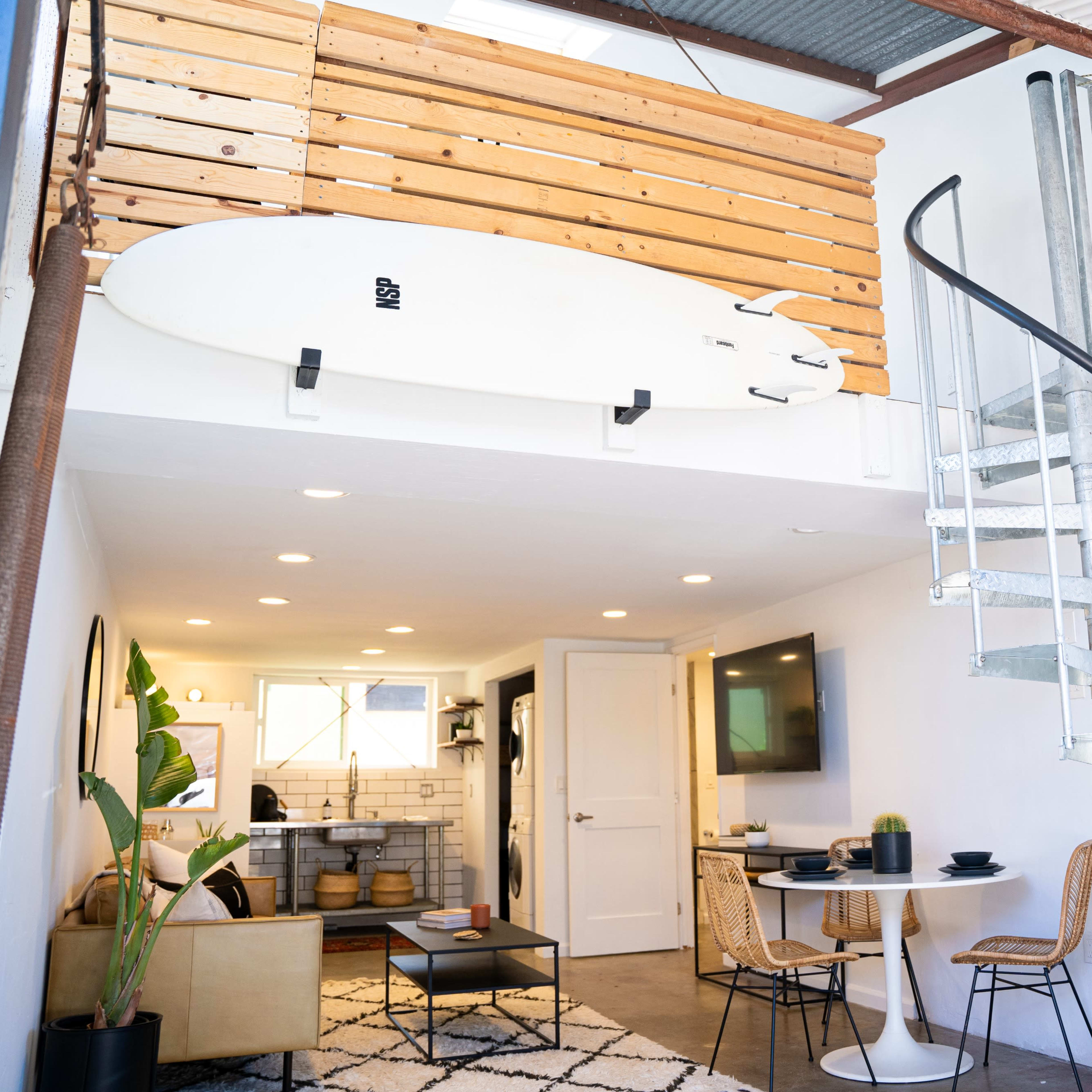A modern, open living space with a spiral staircase, a surfboard mounted on a wooden wall, and a kitchen area visible in the background.