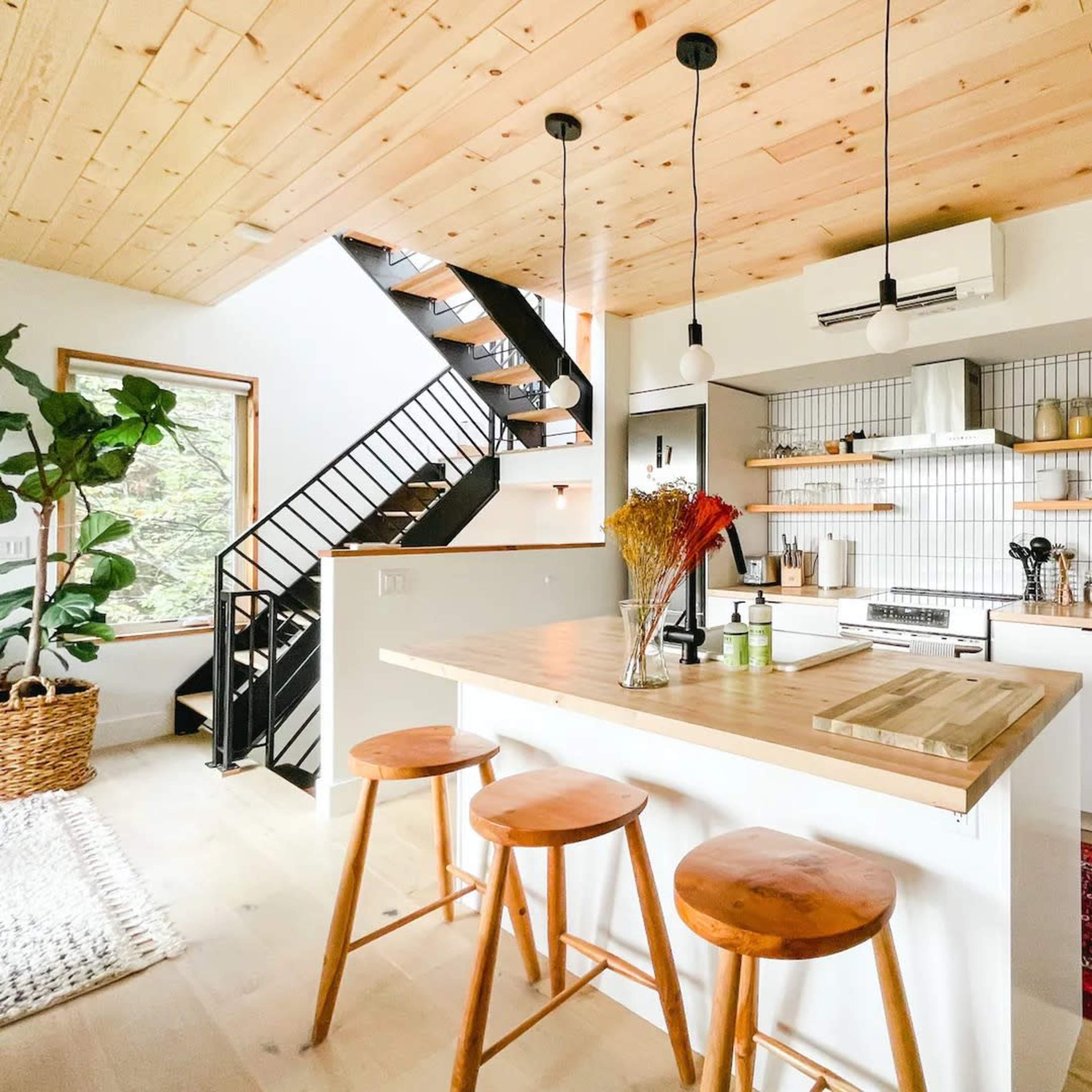 A modern kitchen featuring a wooden countertop with three stools, open shelving with various kitchen items, and a staircase in the background.