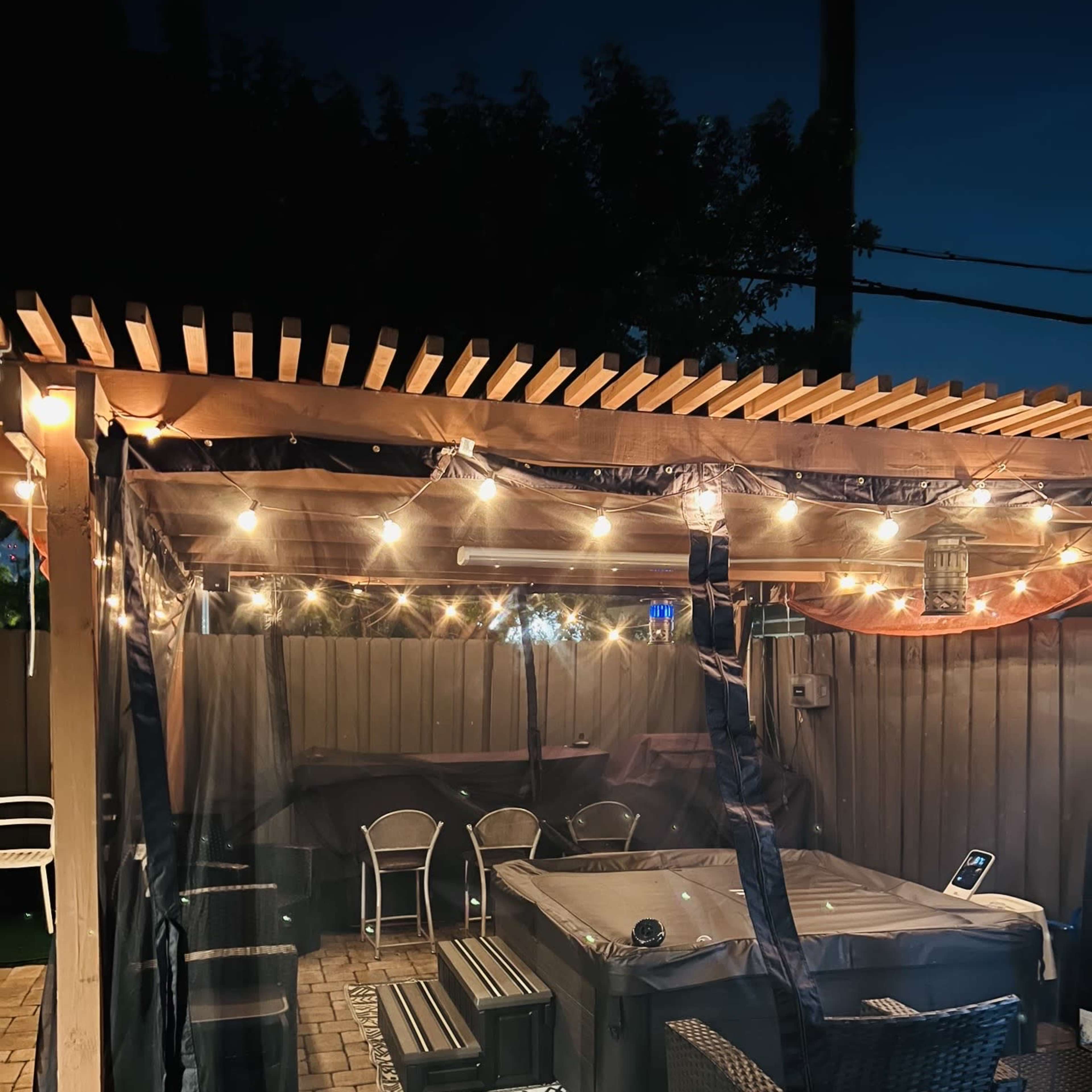 A covered outdoor space with string lights, a hot tub, and seating arrangements surrounded by a netted enclosure.