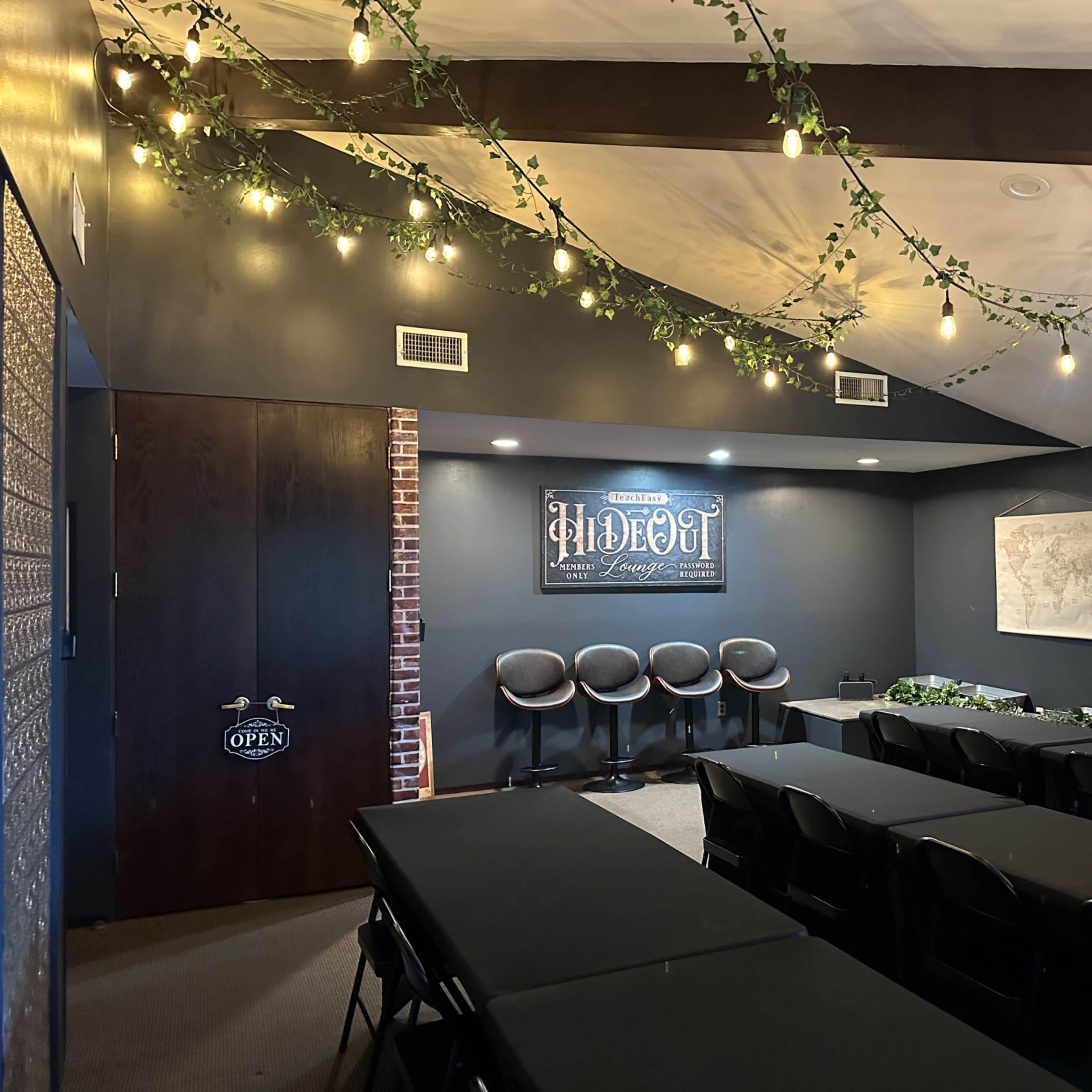 A conference room setup with black tables and chairs, decorated with string lights and greenery, featuring a wall sign that reads "Hideout" and a TV on one side.