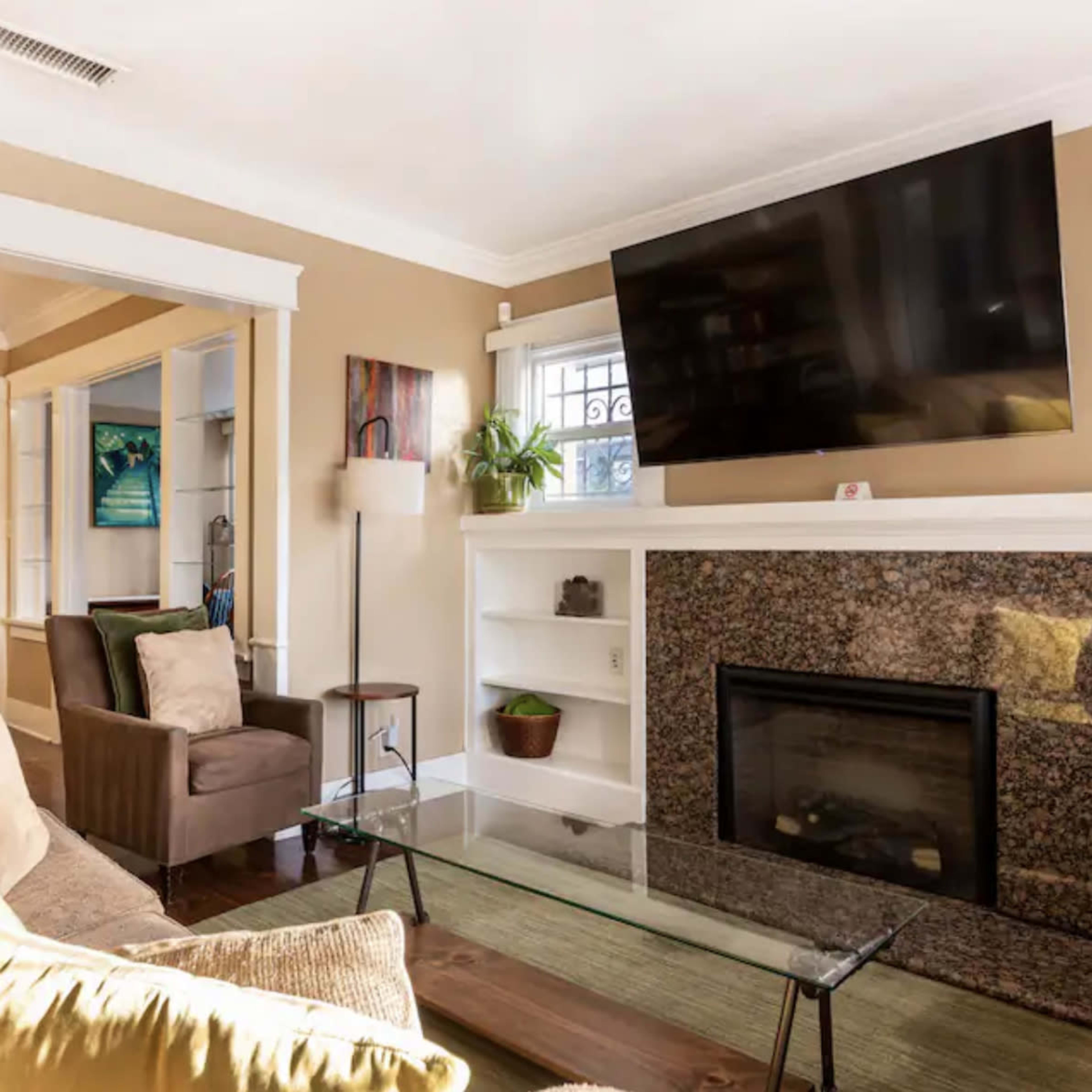 A living room with a sofa, armchair, flat-screen television mounted on the wall, and a stone fireplace beneath it.