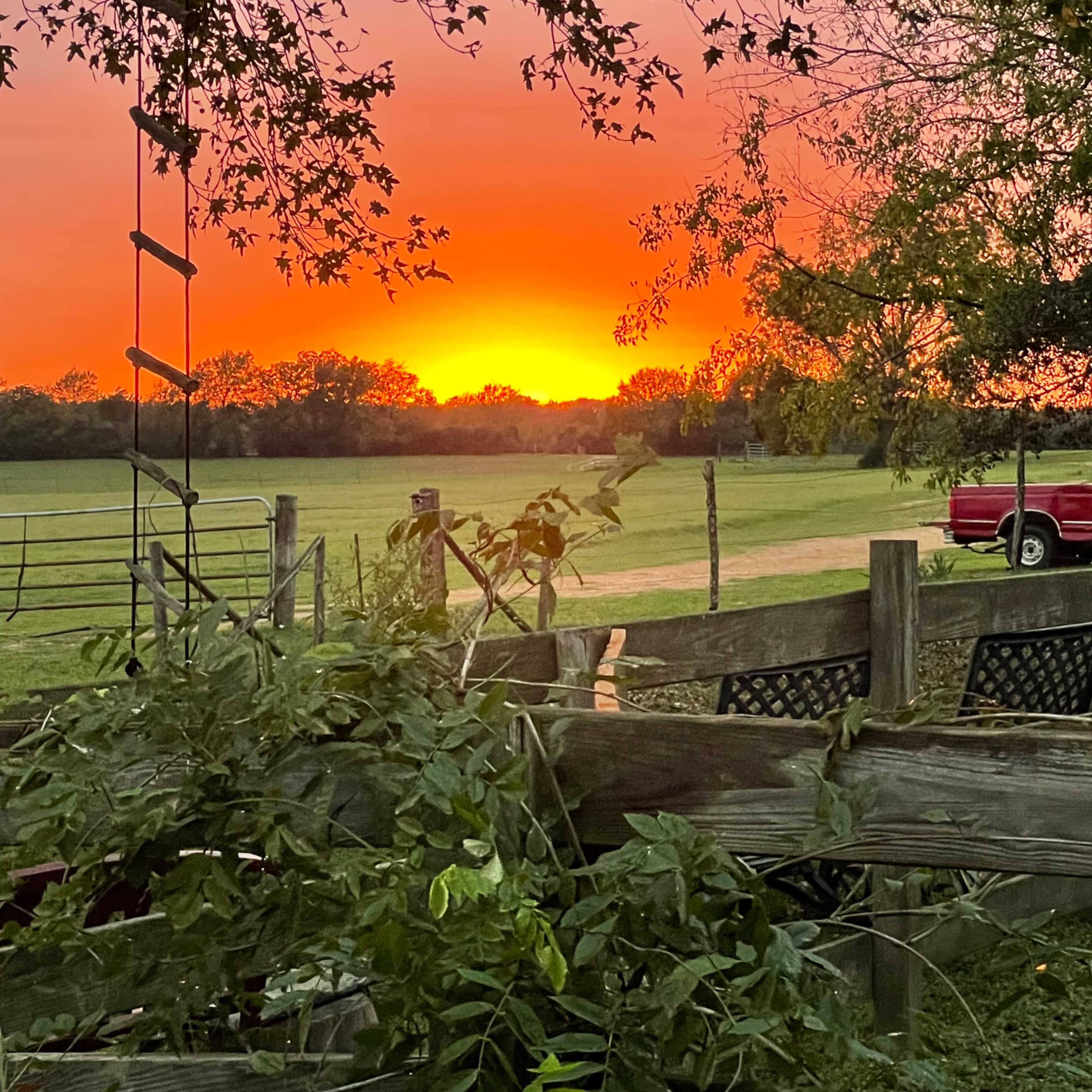 The sun sets behind a fenced pasture, casting a vibrant orange glow in the sky.
