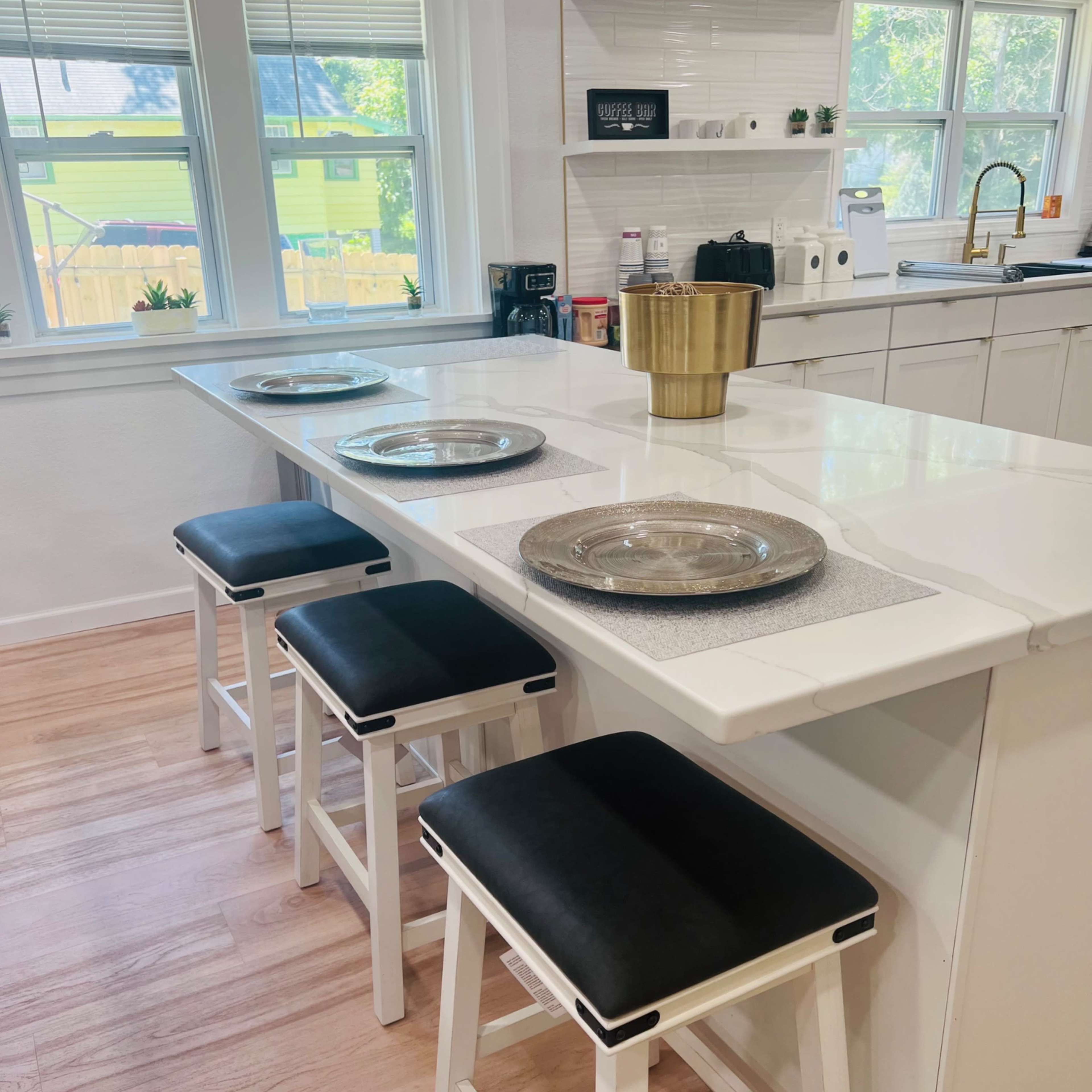 A modern kitchen features a white countertop island with four black and white stools, set with decorative plates and a gold bowl.