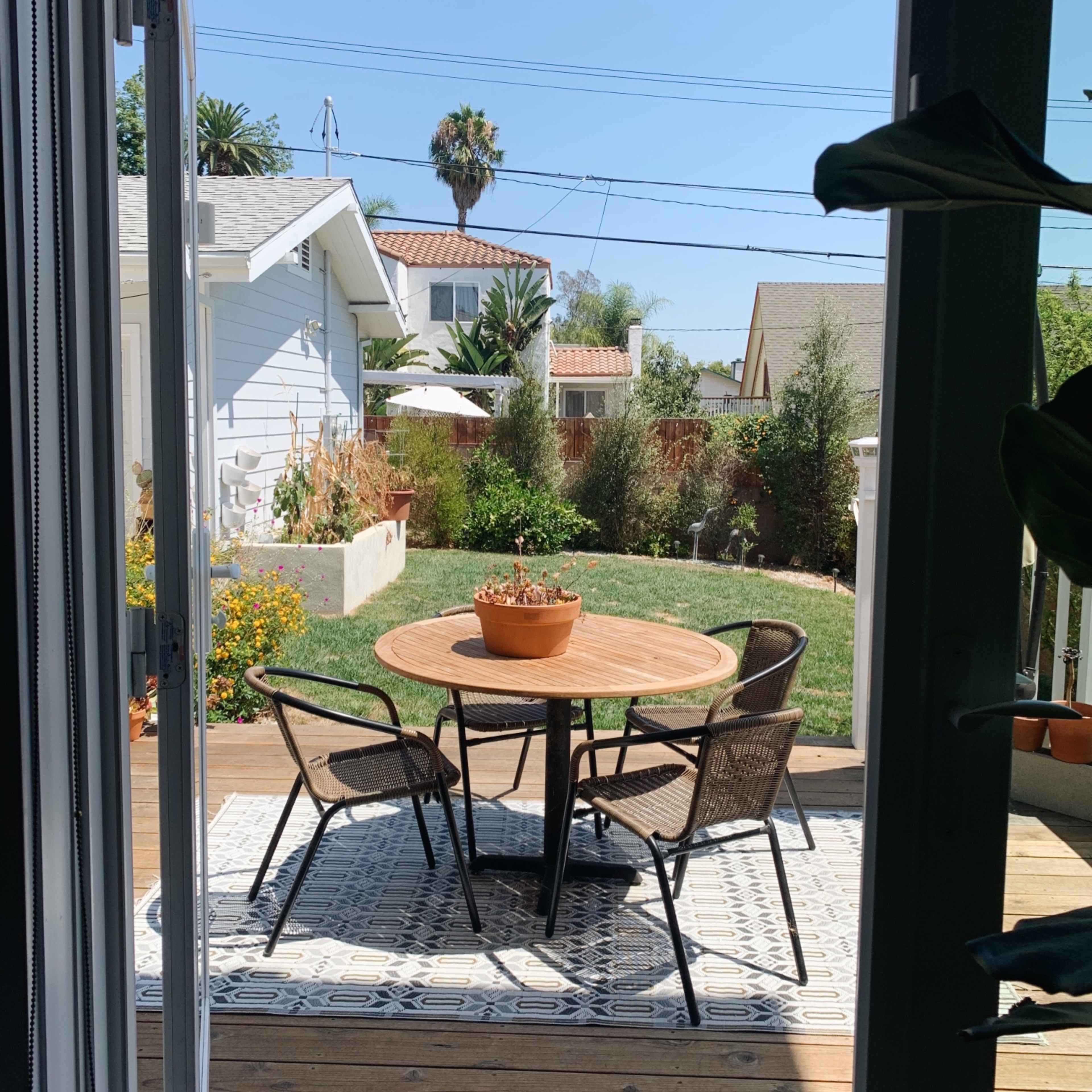 A round wooden table with four chairs sits on a patio overlooking a landscaped backyard with greenery and a palm tree.
