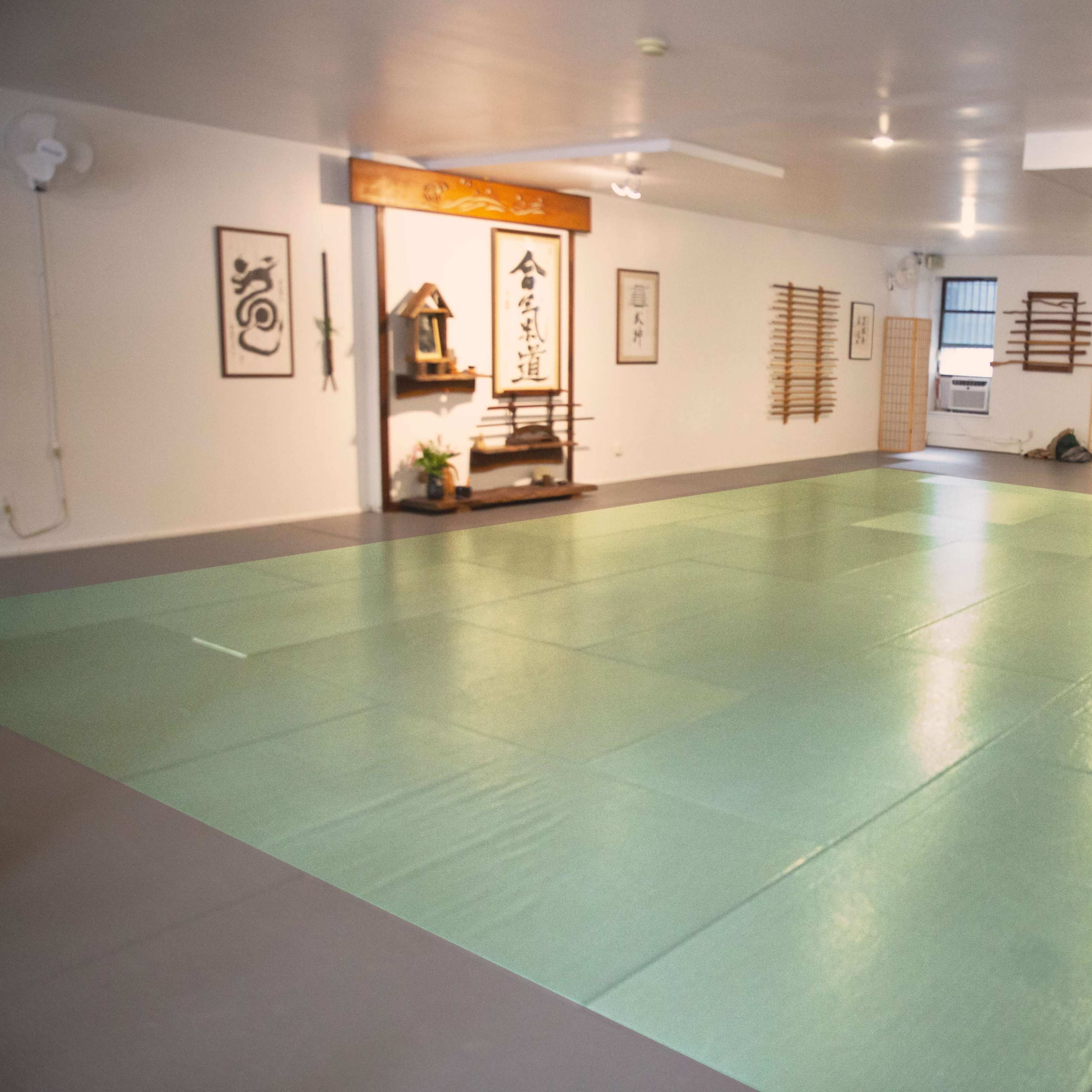 A spacious martial arts dojo with green mats, wooden wall decorations, and a traditional display area.