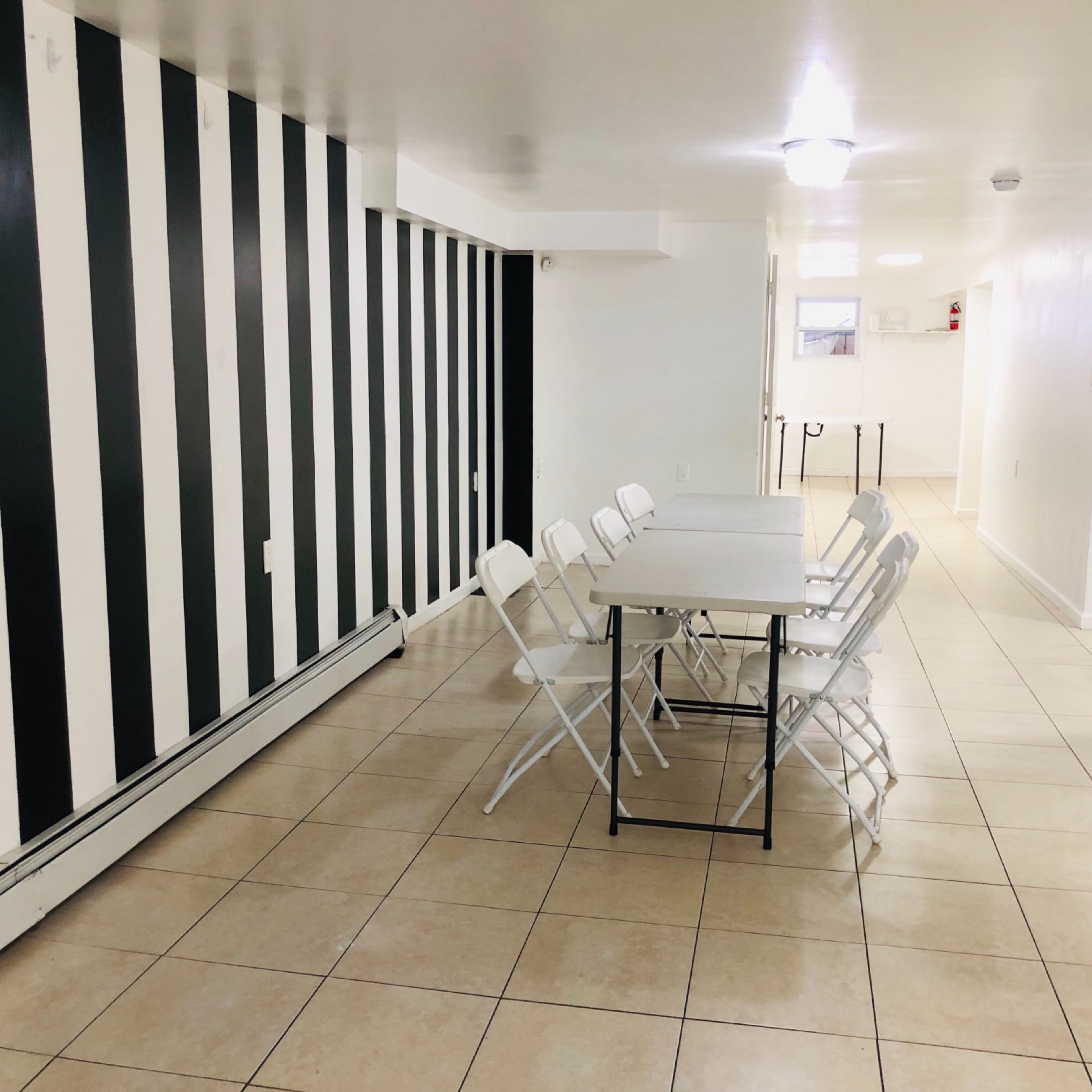 A row of white folding chairs surrounds a rectangular table in a spacious, tiled room with black and white striped walls.