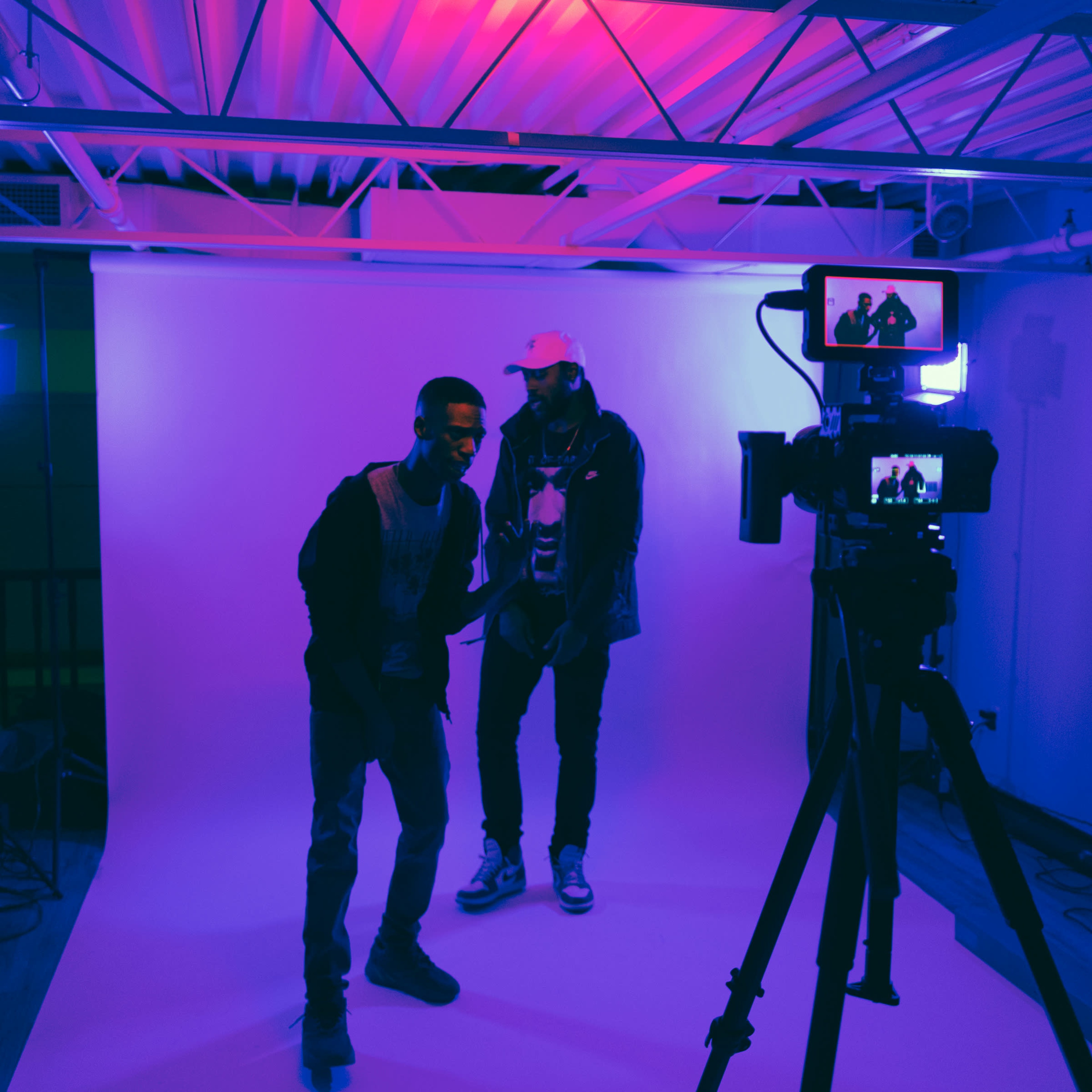 Two men are posing in front of a camera with a white backdrop and colorful lighting.