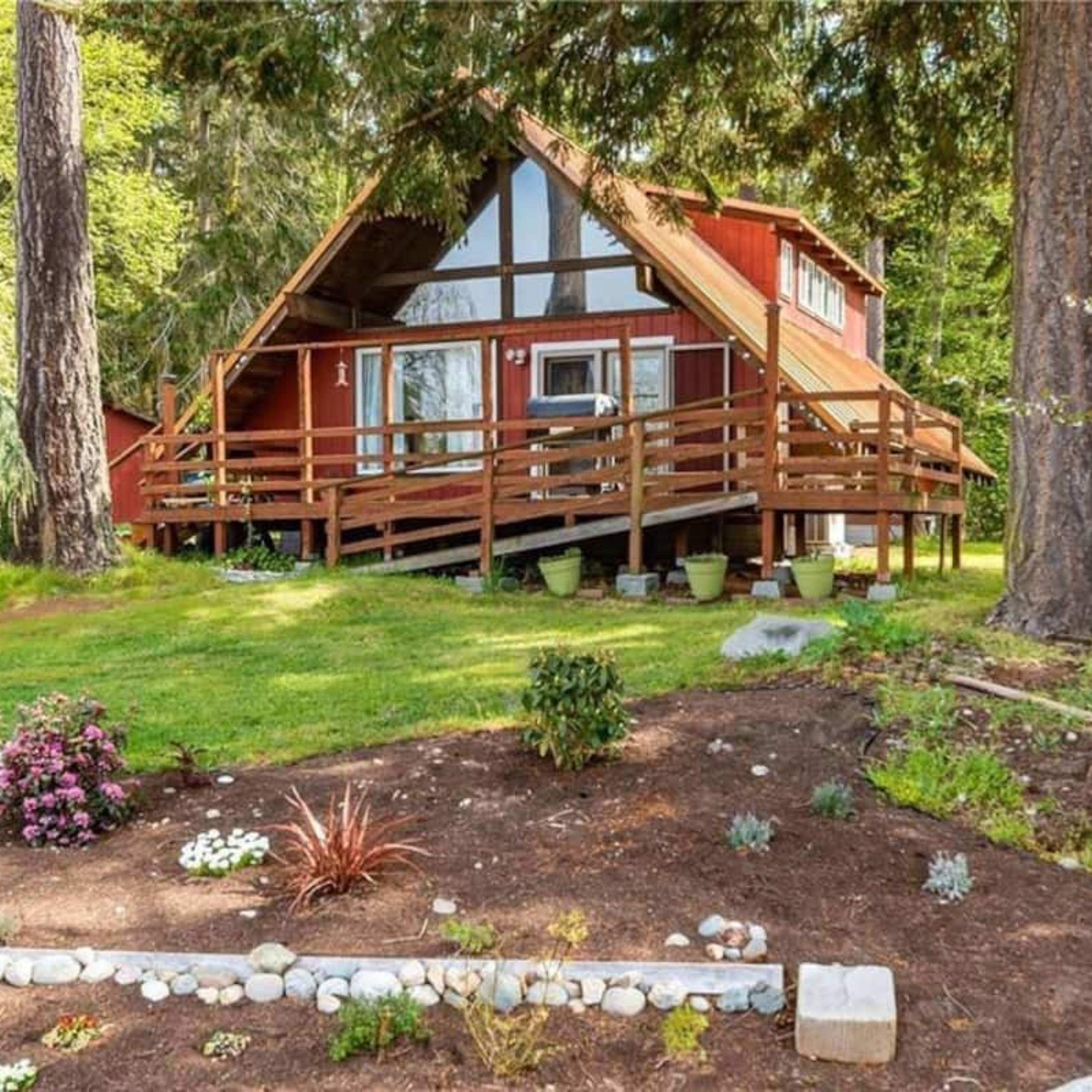 A red A-frame house with a wooden deck sits on a grassy area, surrounded by trees and flower beds.