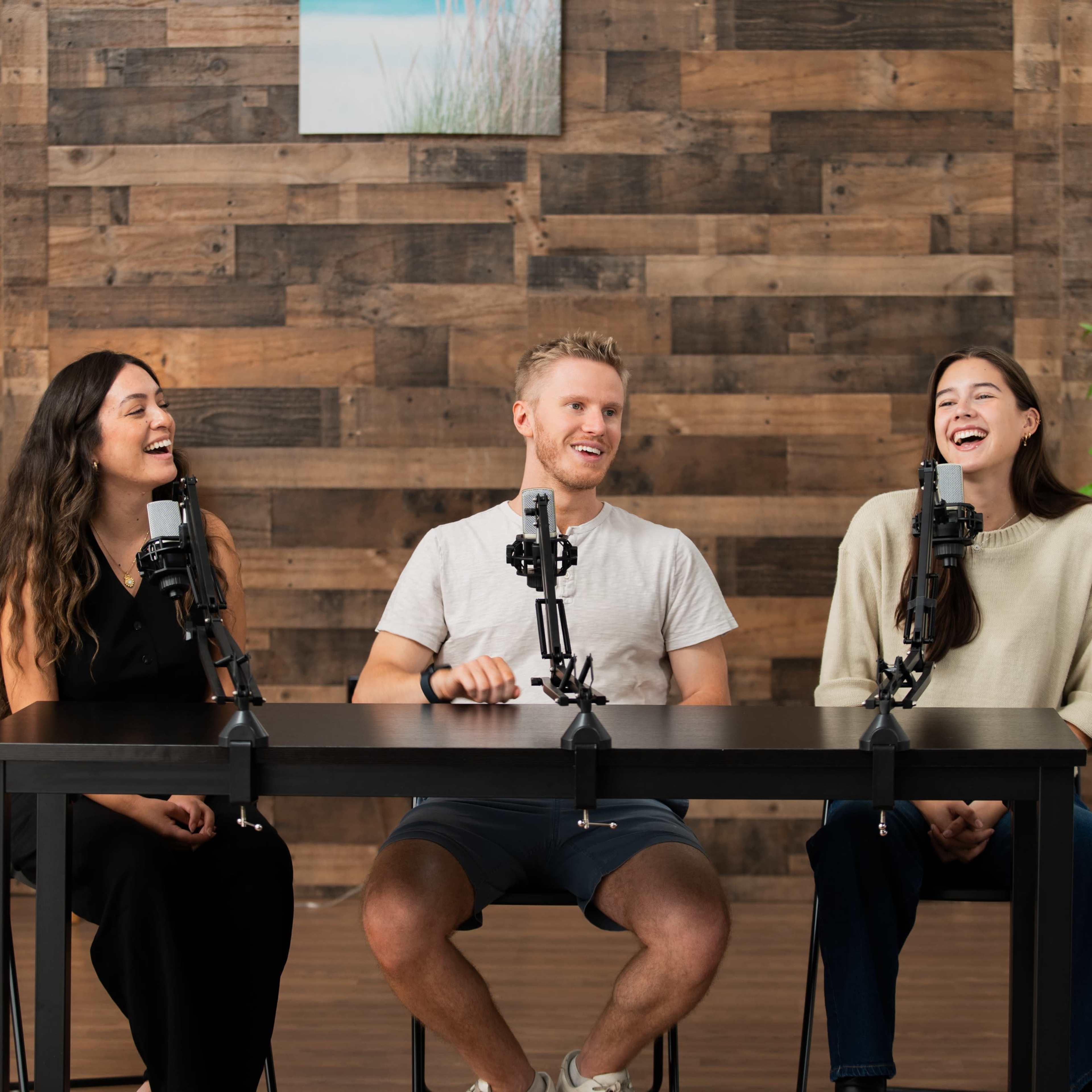Three people are seated at a table with microphones in front of them, smiling and laughing in a wooden-paneled studio.