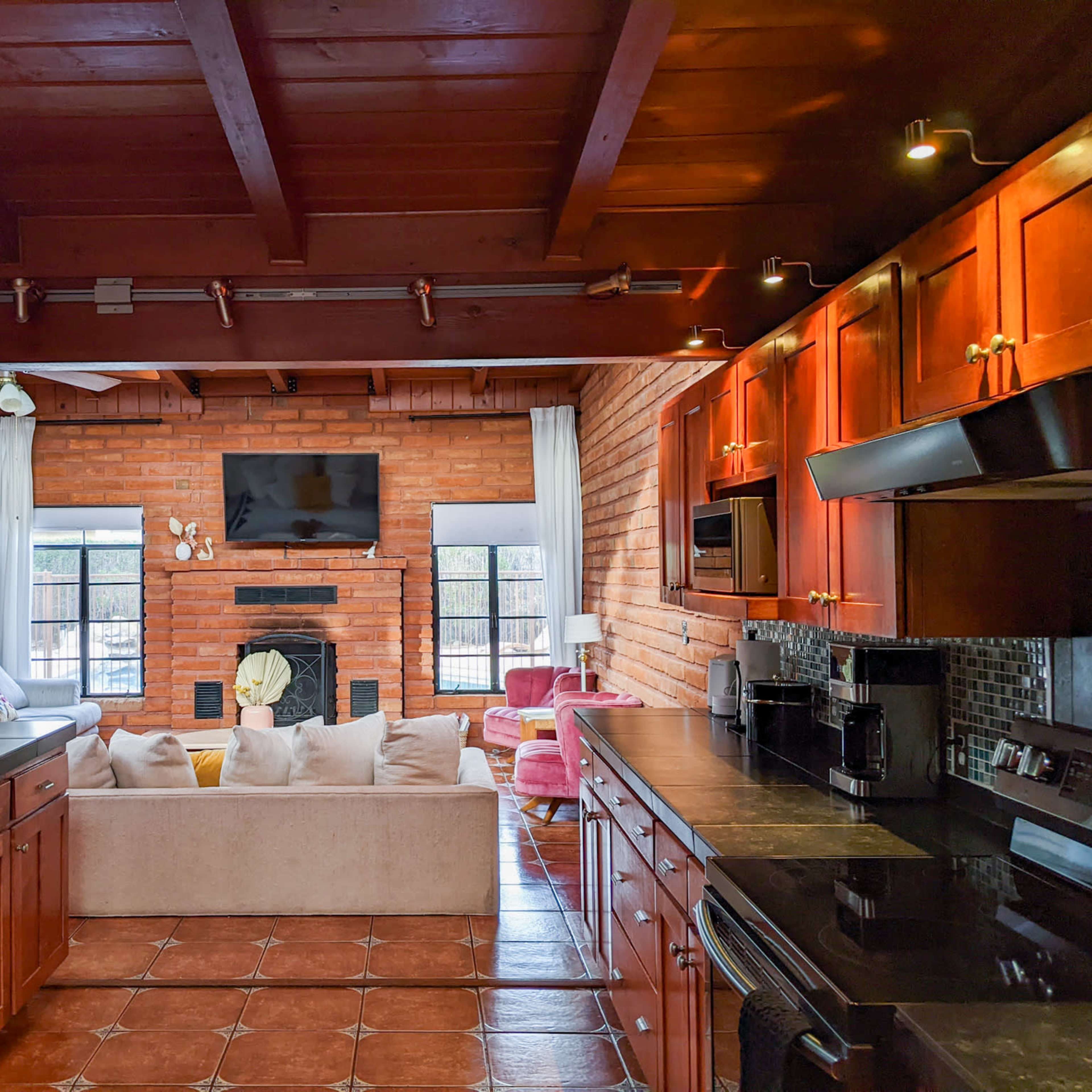 A cozy living room with a brick wall, large windows, and a kitchen featuring dark wooden cabinets and black countertops.