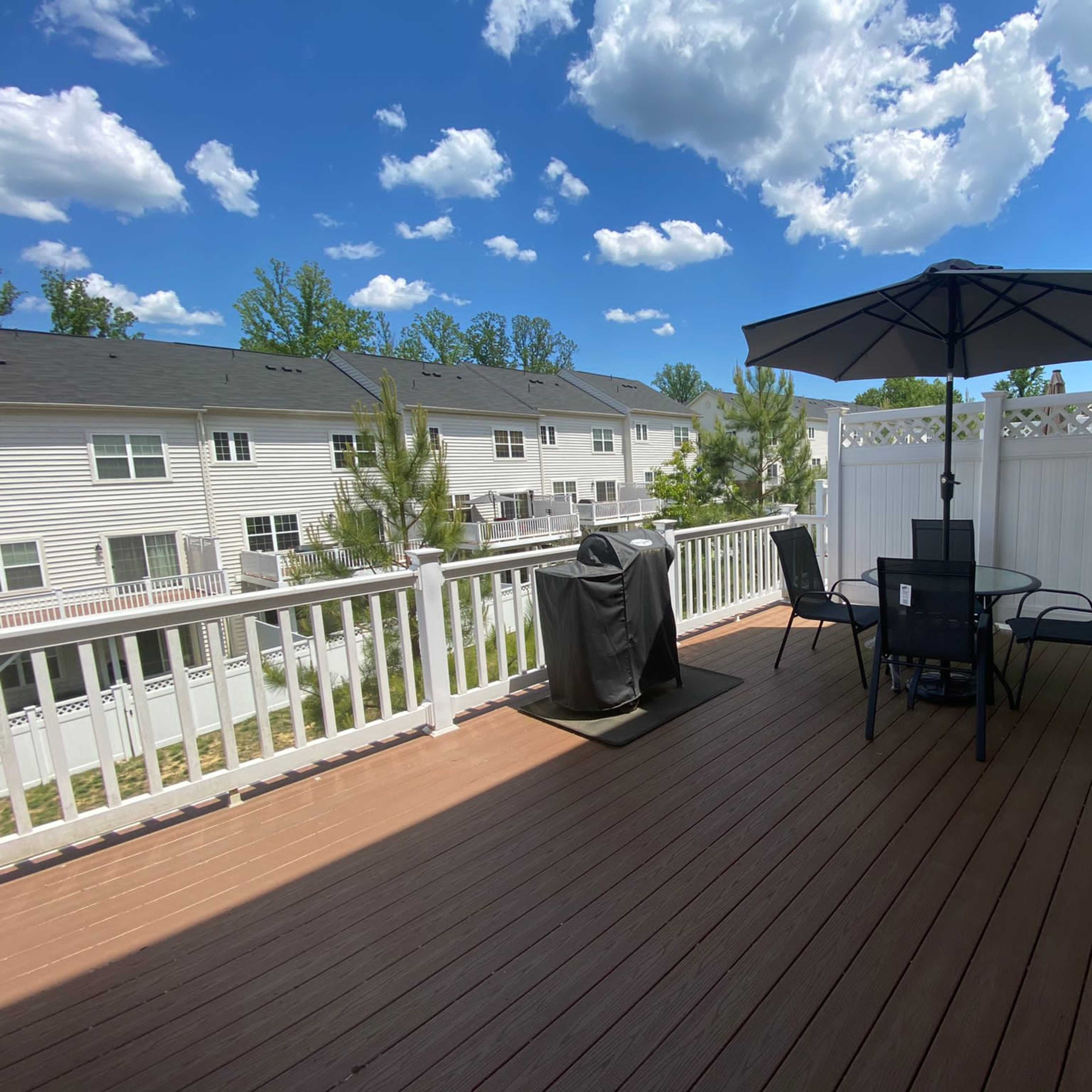 A wooden deck with a table and chairs under an umbrella, overlooking a row of houses and a blue sky with clouds.