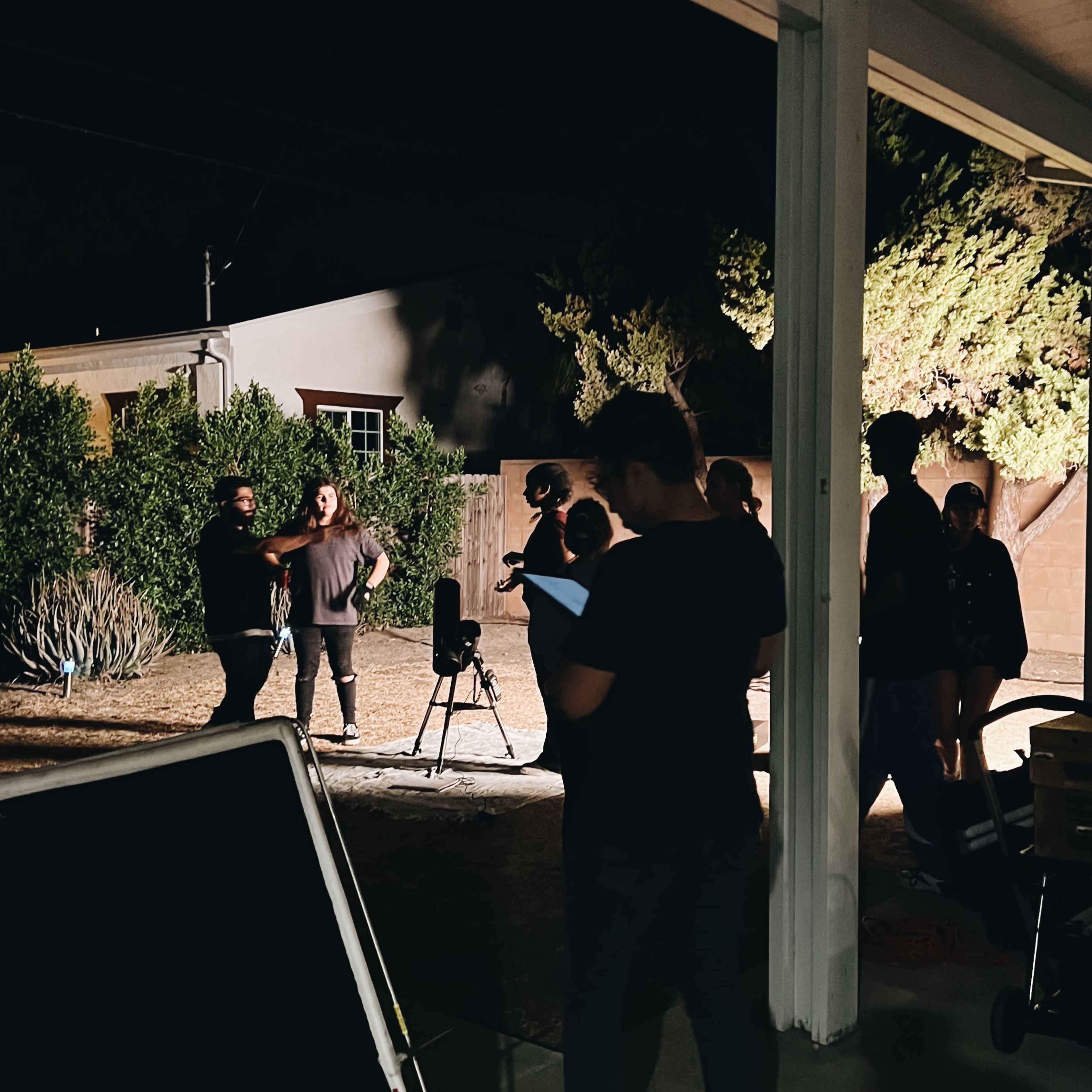 A group of people is filming a scene in a backyard at night, illuminated by artificial lights.
