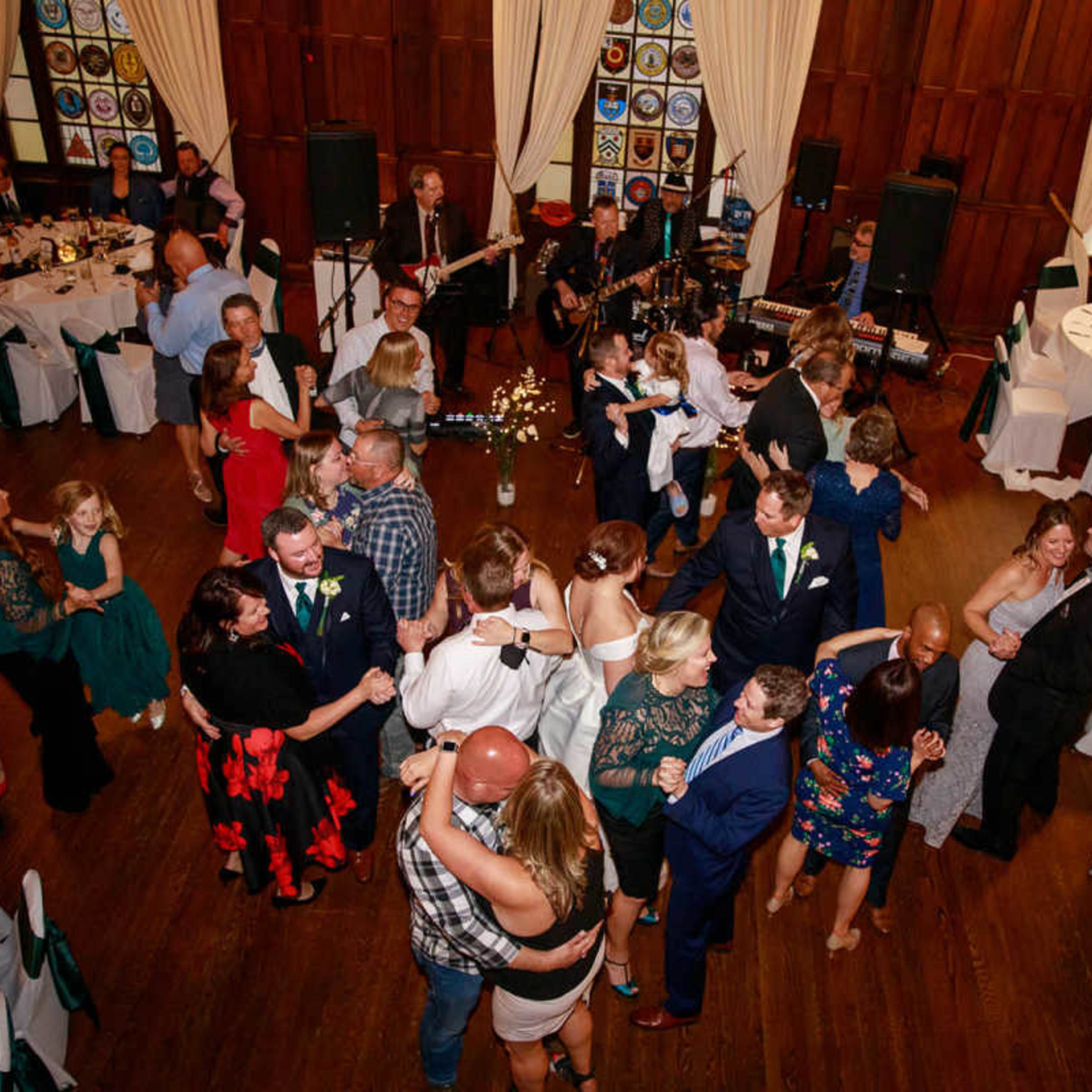 A spacious banquet hall filled with guests dancing and mingling, with tables set around the dance floor and a band performing in the background.