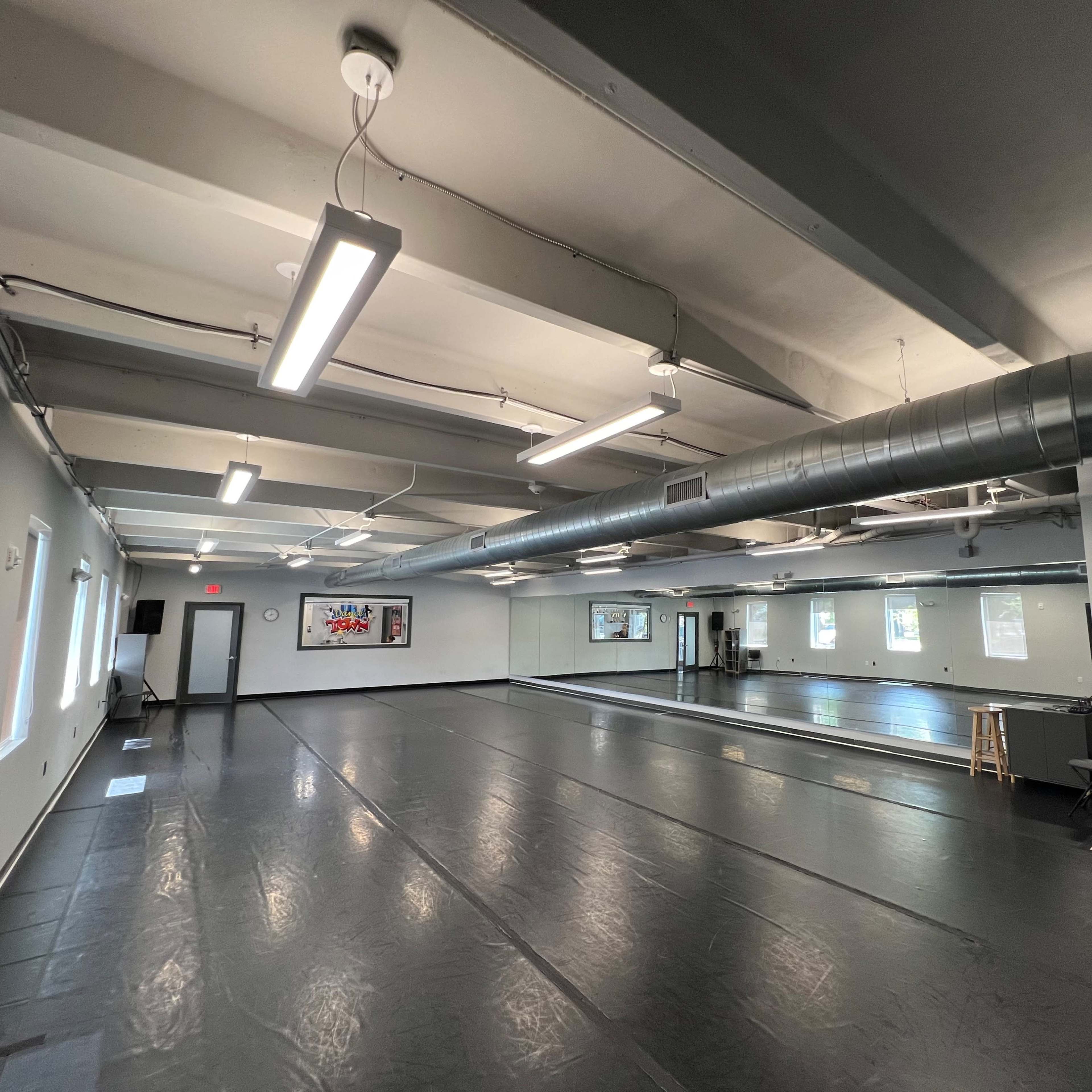 An empty dance studio with mirrored walls, a black floor, and overhead lighting.