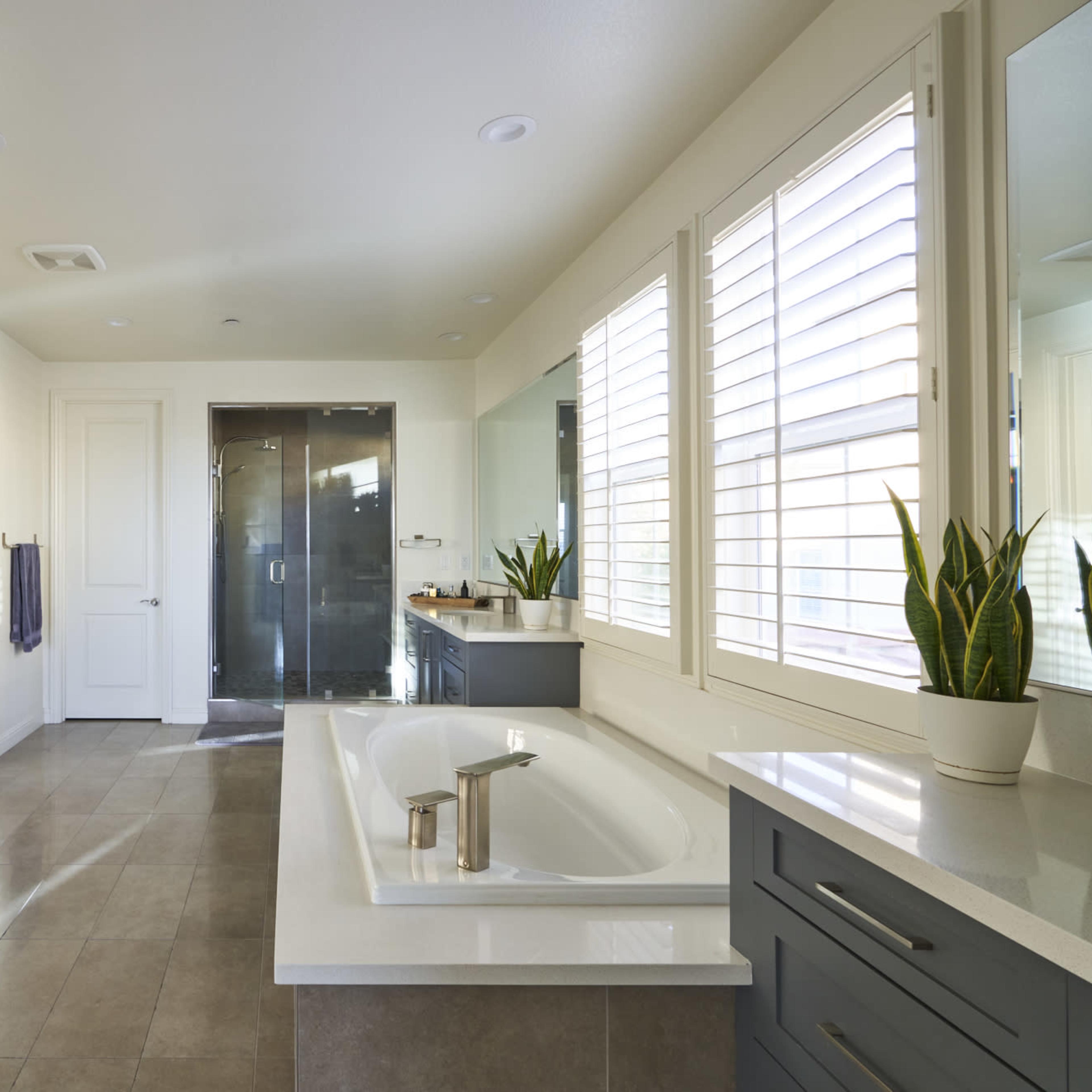 The bathroom features a freestanding bathtub, dual sinks with gray cabinetry, and a glass-enclosed shower, illuminated by natural light from the windows.