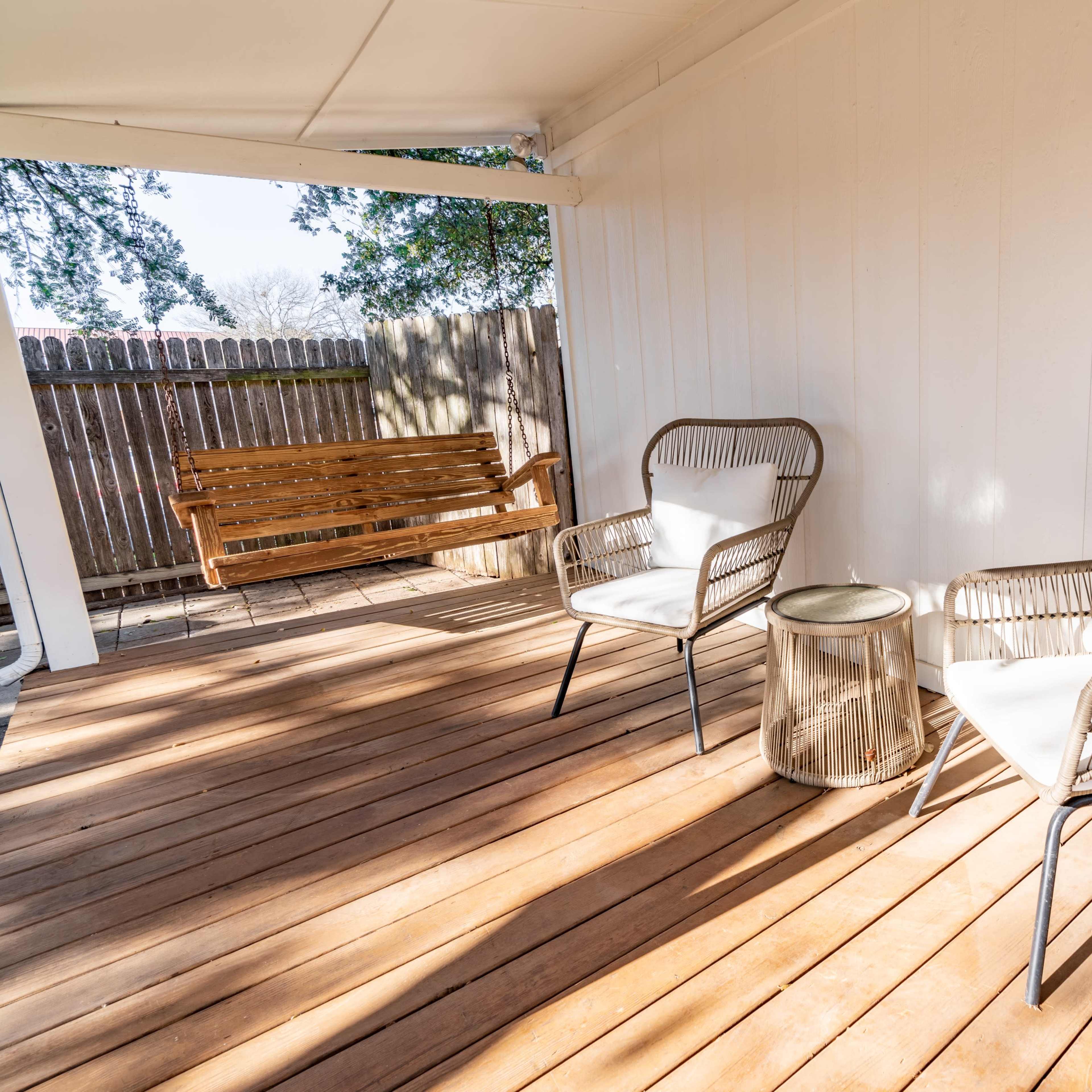 A patio area with two wicker chairs, a small table, and a wooden bench against a wooden fence.