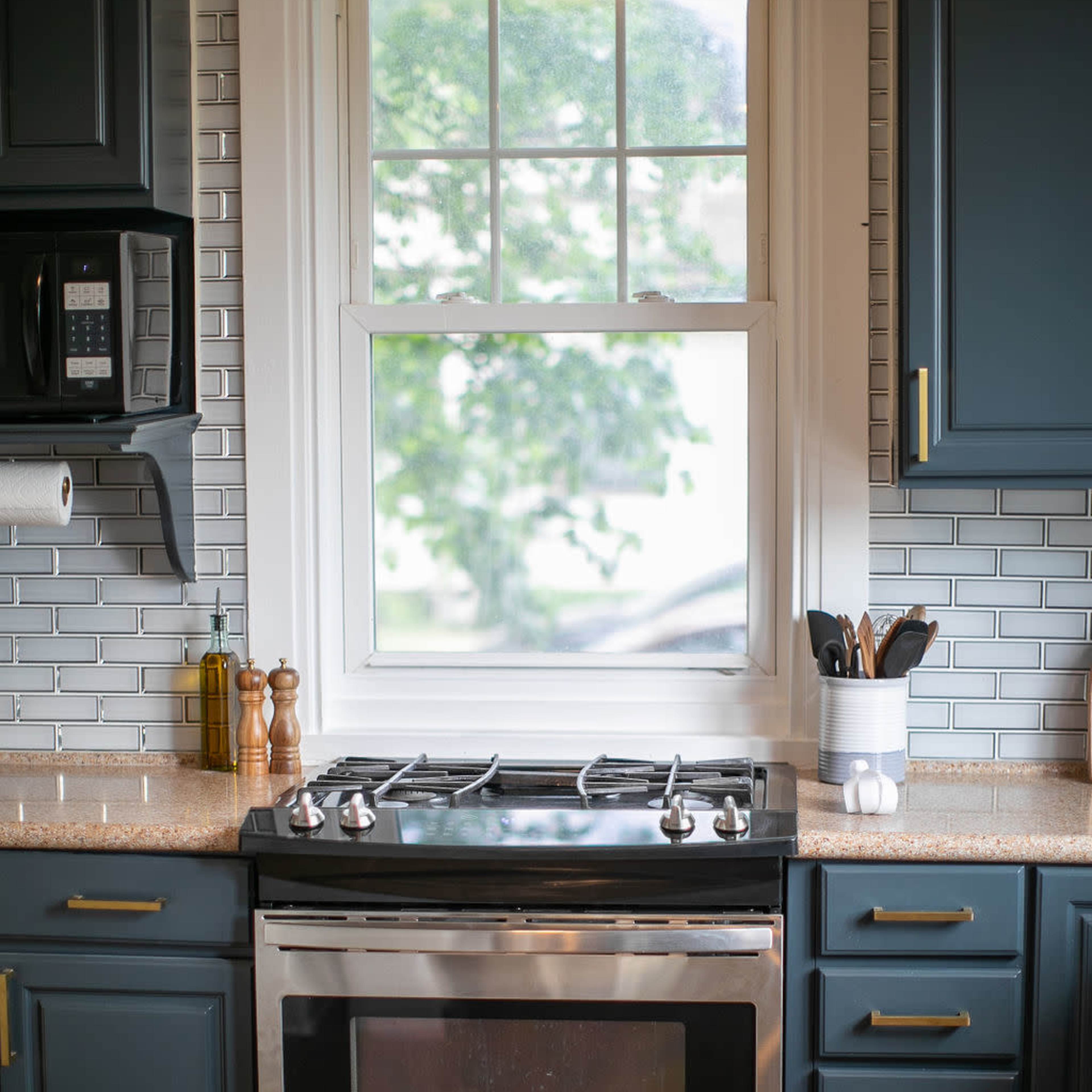 A stainless steel stove is placed beneath a window in a kitchen with dark blue cabinetry and a tiled backsplash.