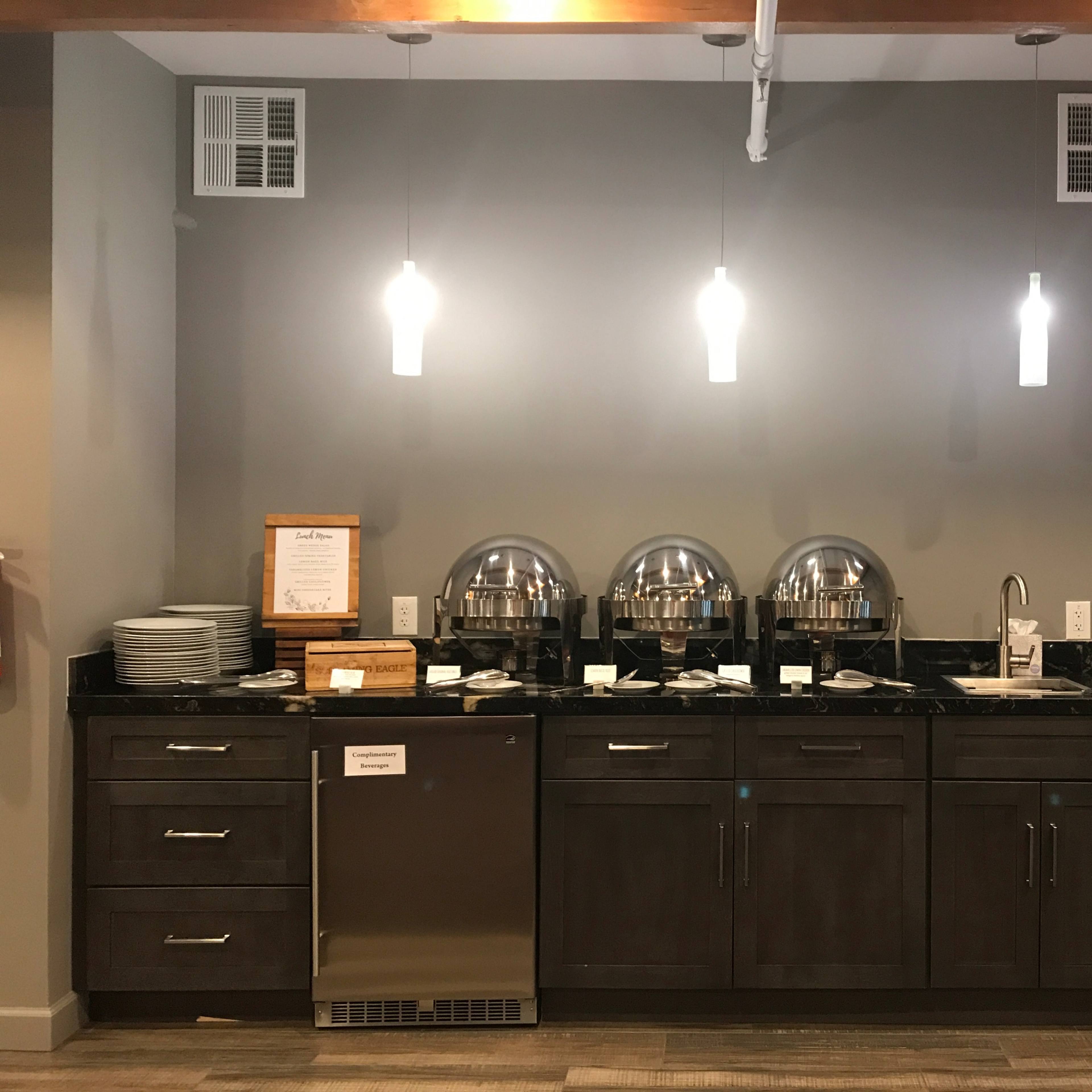 A modern buffet station with a dark countertop, several chafing dishes, plates, utensils, and a stainless-steel refrigerator against a gray wall.