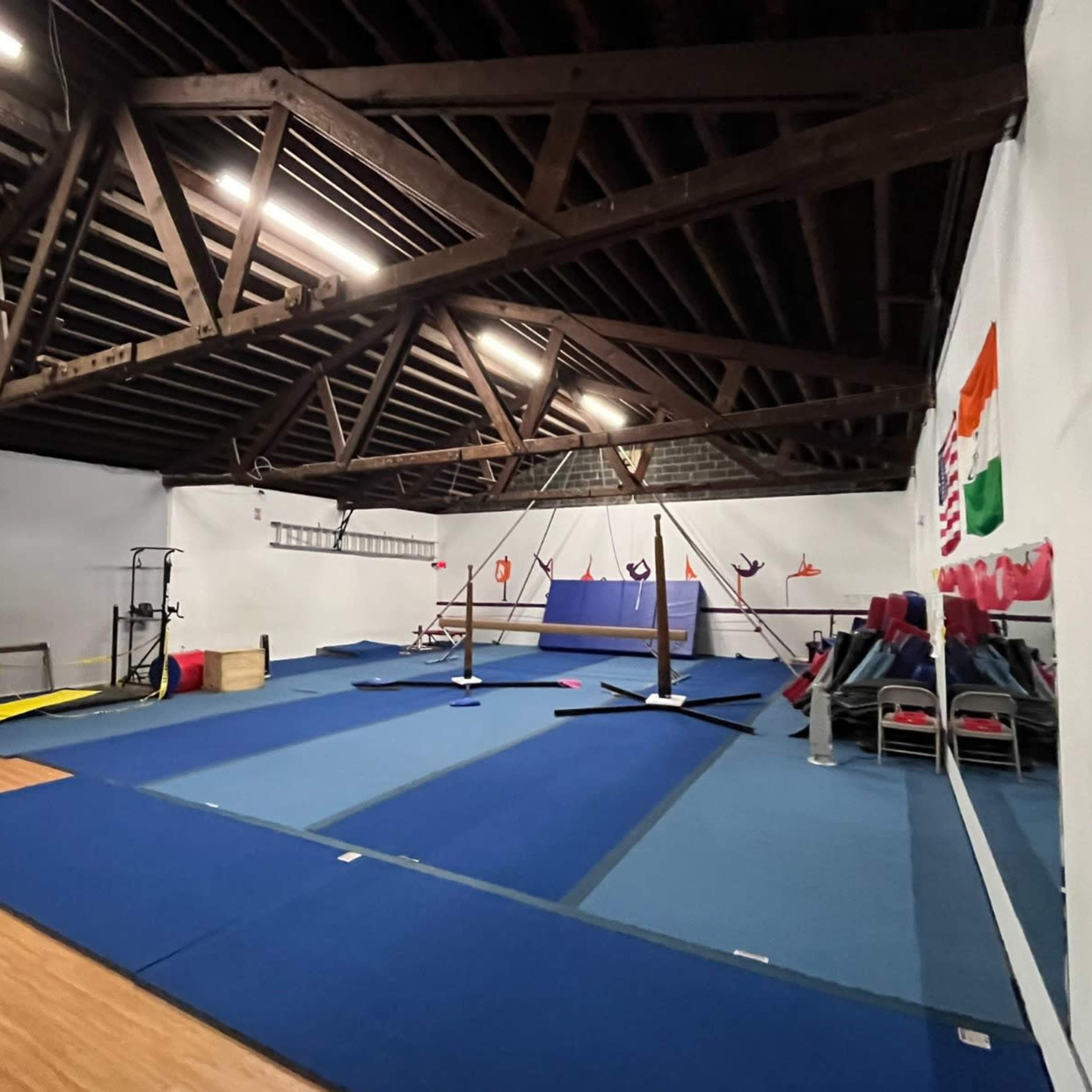 A spacious gymnasium with wooden beams, featuring gymnastics equipment and blue mats spread across the floor.