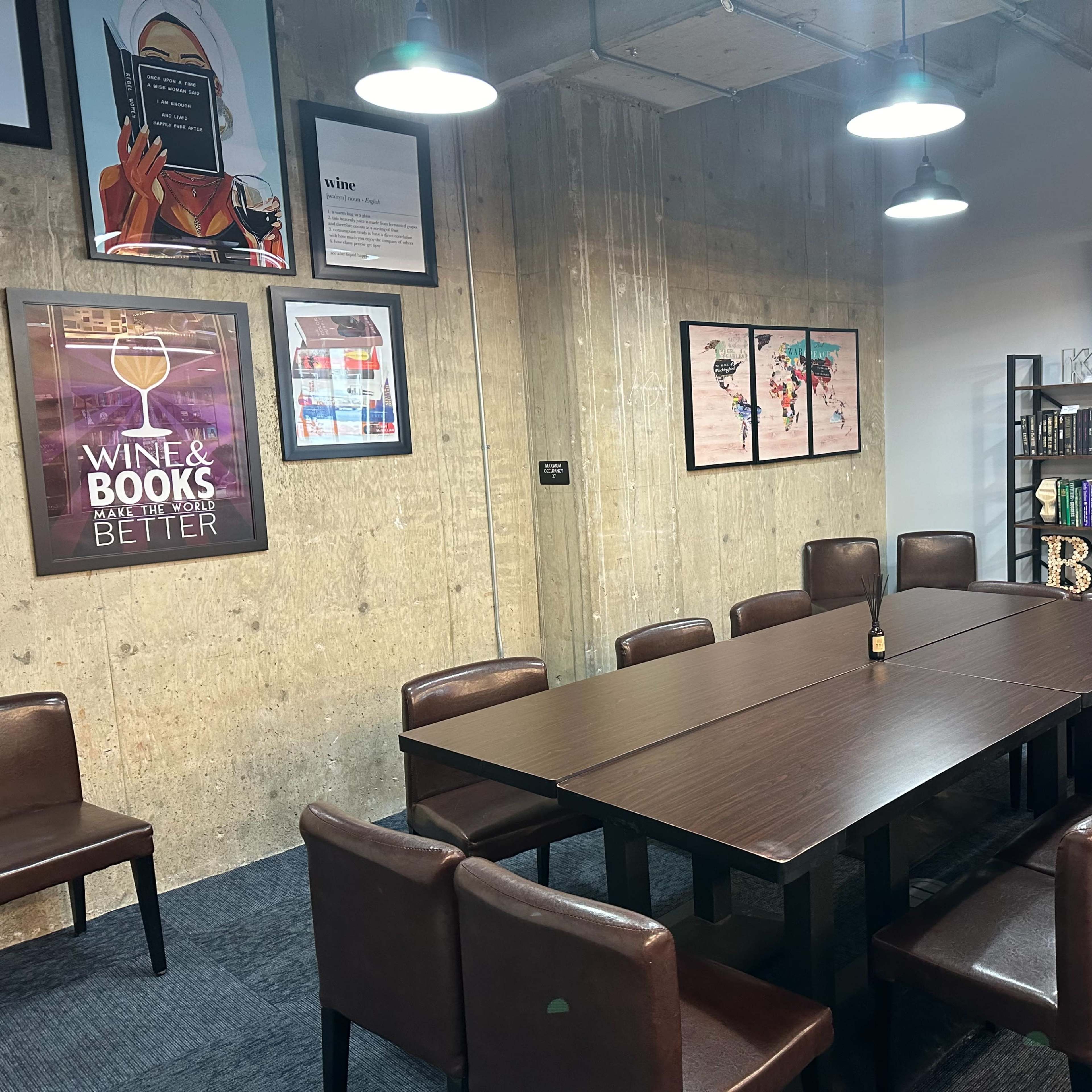 A conference room with a large table surrounded by brown chairs, featuring colorful artwork and shelves filled with books against a concrete wall.