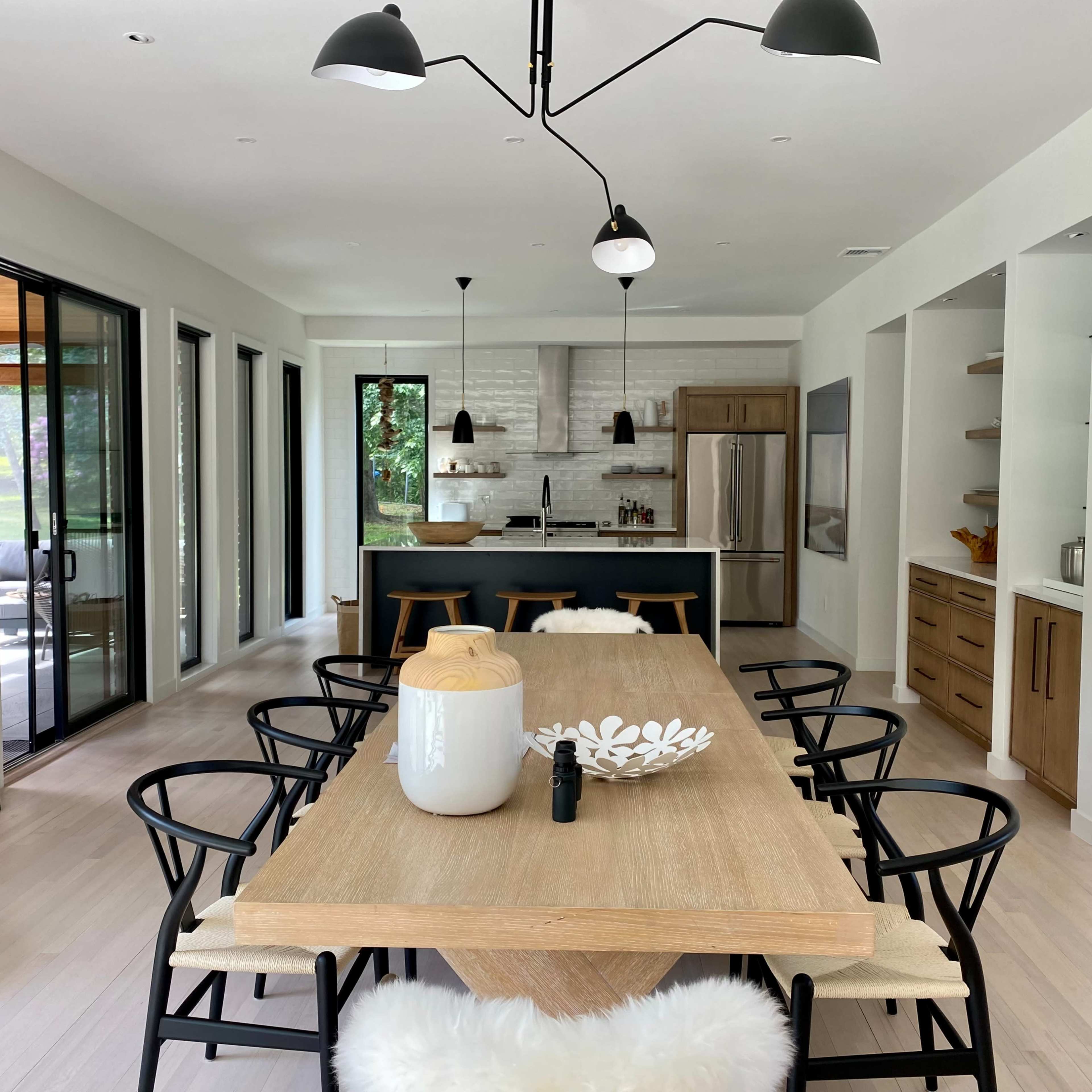A modern dining area features a long wooden table surrounded by black chairs, leading to an open kitchen with a large island and stainless steel appliances.