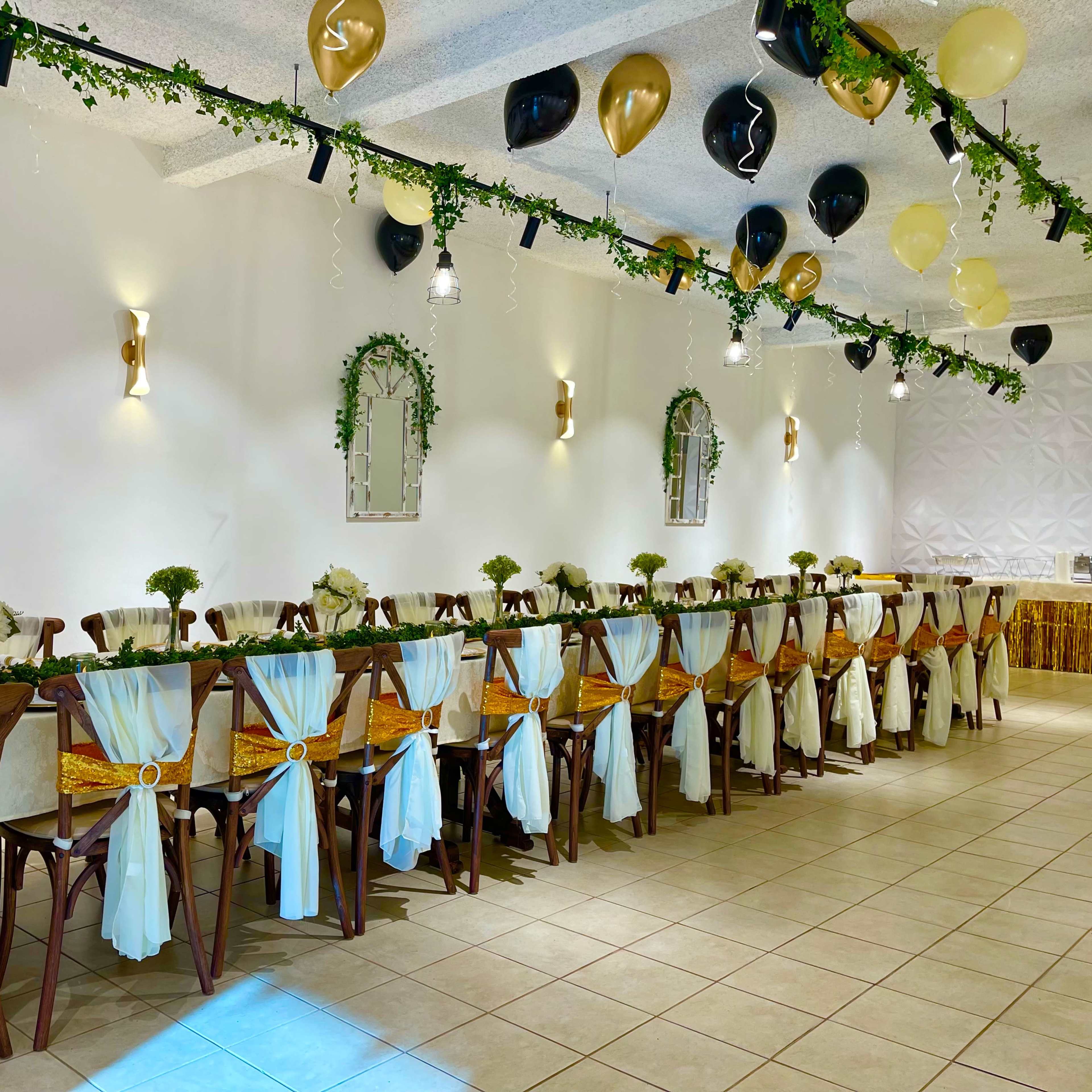 A long, elegantly set dining table is adorned with white and gold decorations, surrounded by wooden chairs and decorated with floral centerpieces, in a well-lit event space.