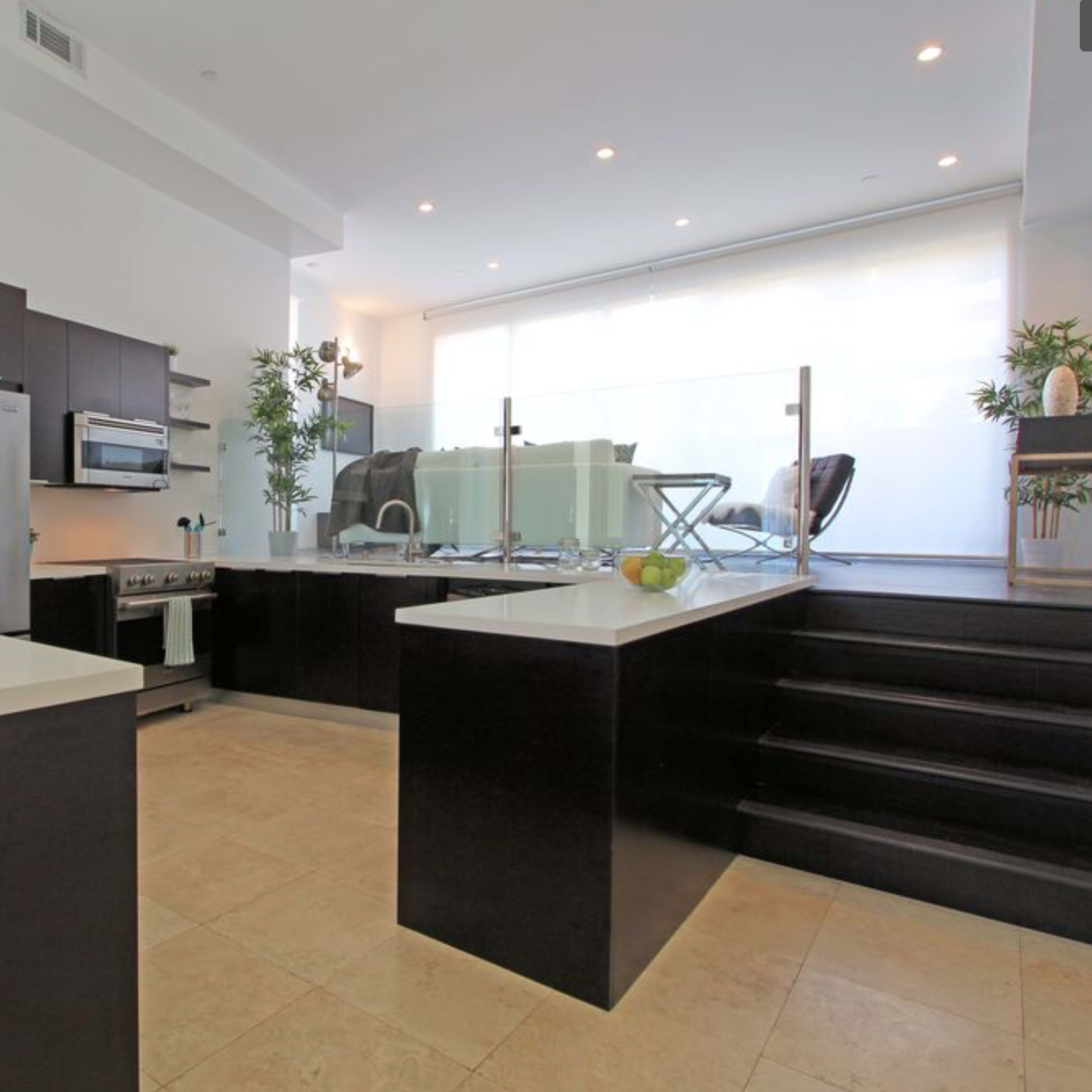 A modern kitchen with dark cabinetry, white countertops, and a glass partition leading to a living area with steps.