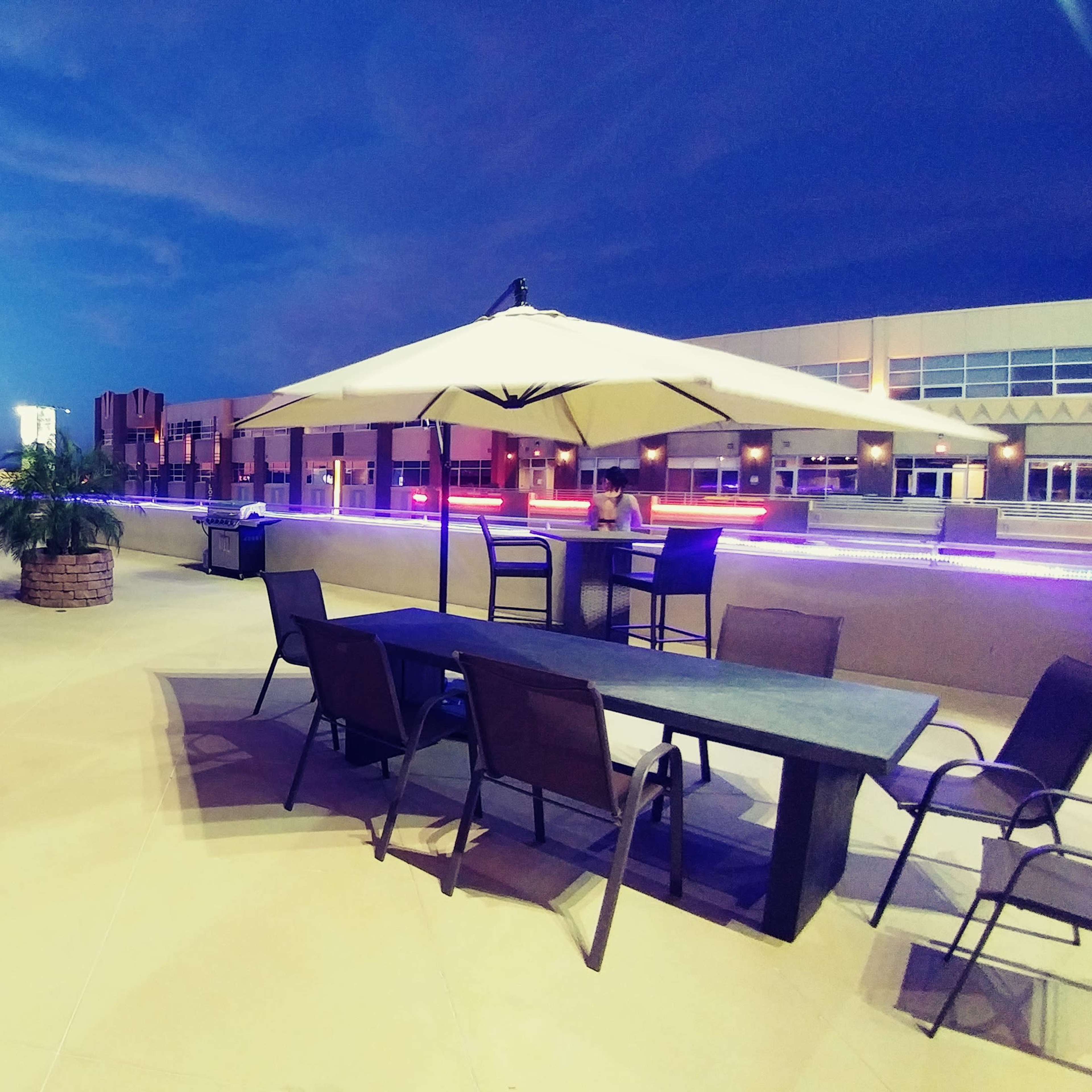A rooftop lounge area with patio furniture, palm plants, and umbrellas under a twilight sky.