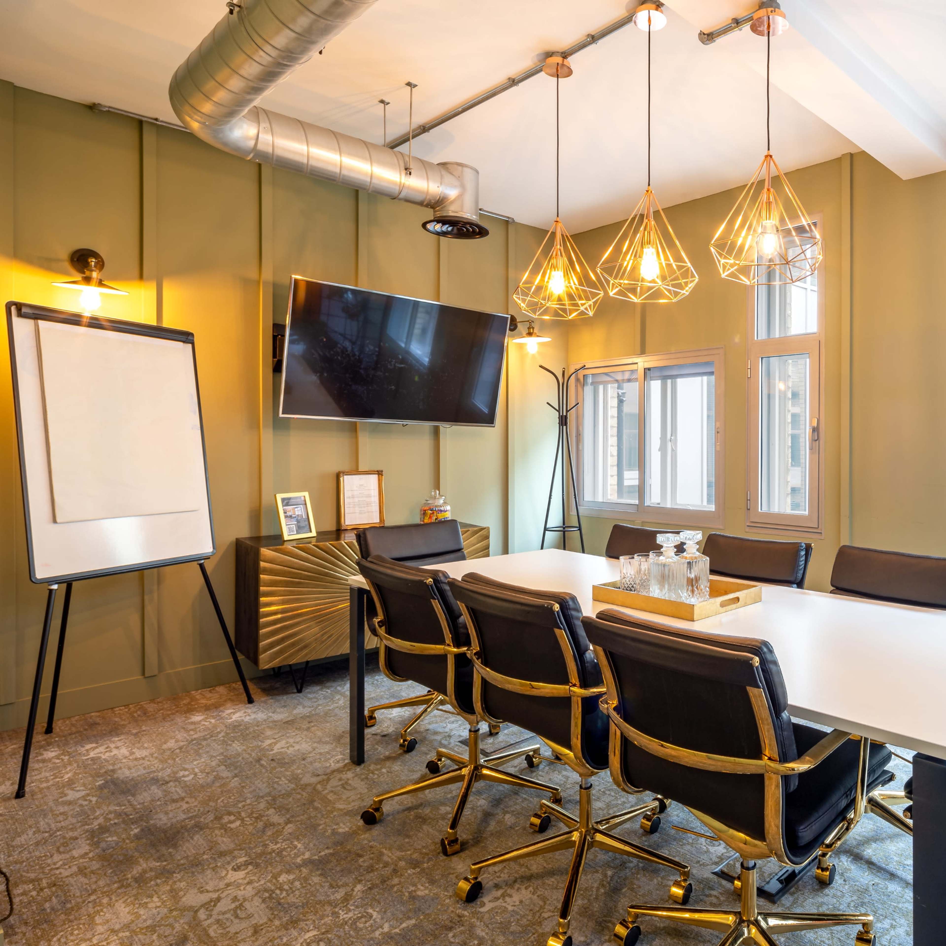 A modern conference room features a long table surrounded by black and gold chairs, a television mounted on the wall, and three pendant lights above.