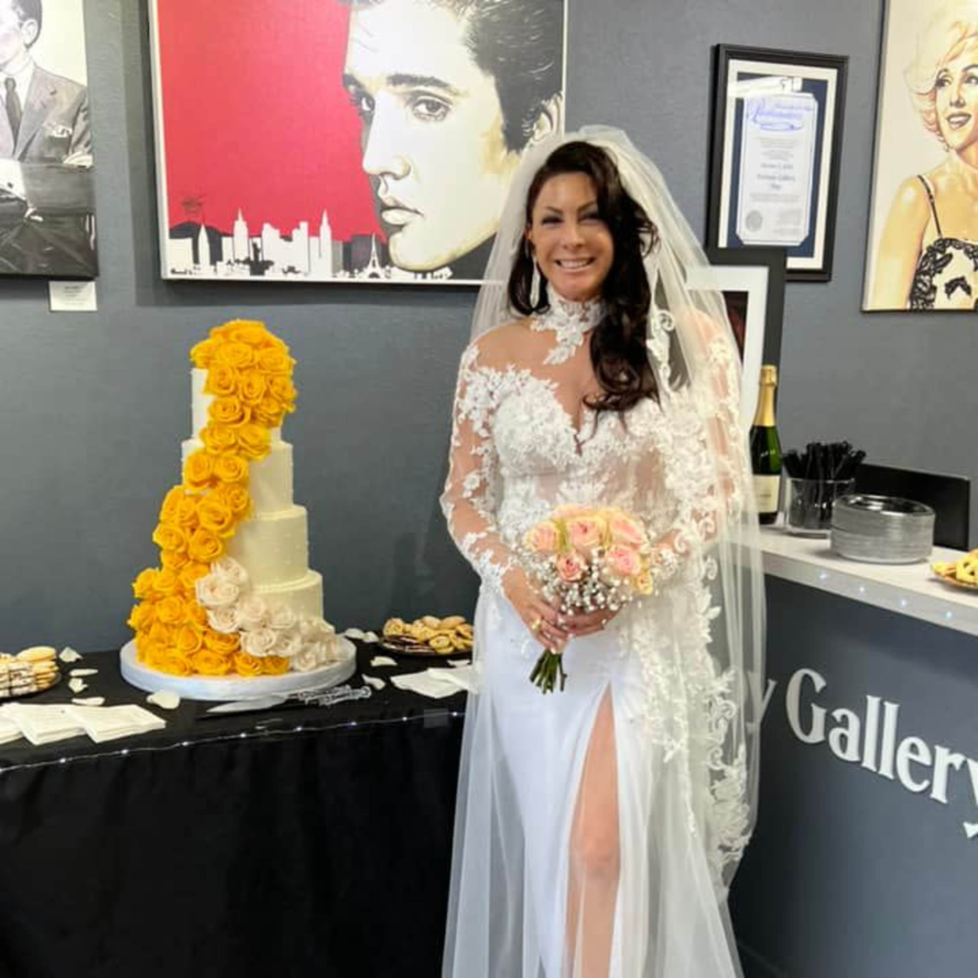 A bride in a white gown stands next to a cake adorned with yellow roses and a table of refreshments.