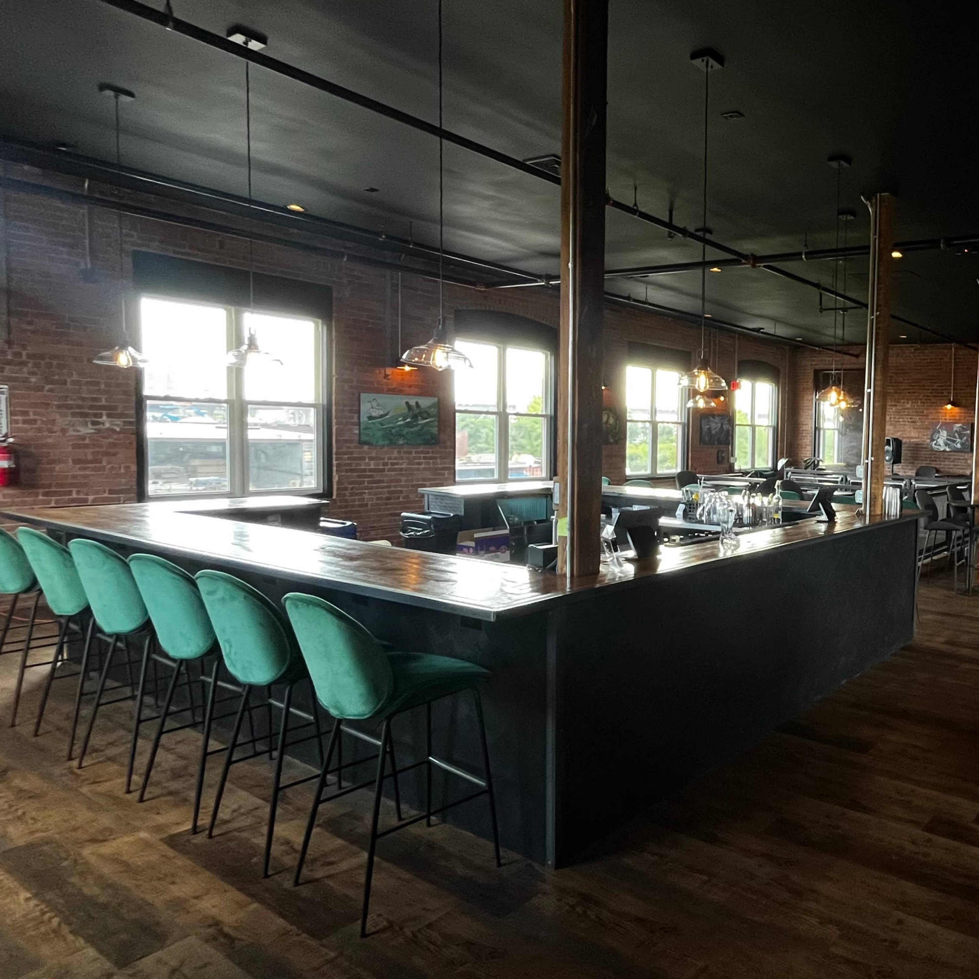 A modern bar area with a long counter, several high stools, and seating tables in a space featuring brick walls and large windows.