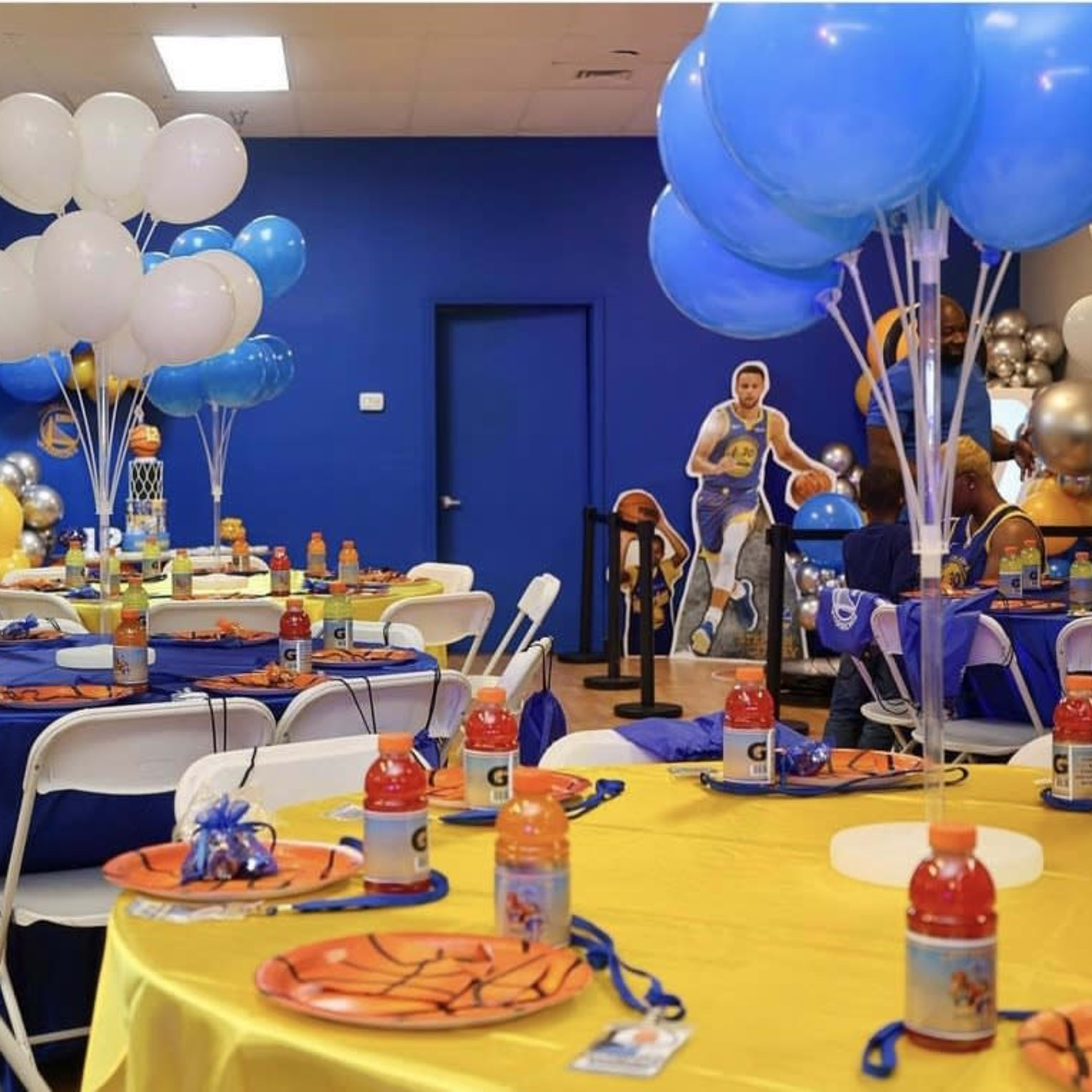 A basketball-themed party setup with tables decorated in blue and yellow, featuring themed plates, Gatorade bottles, and balloons.