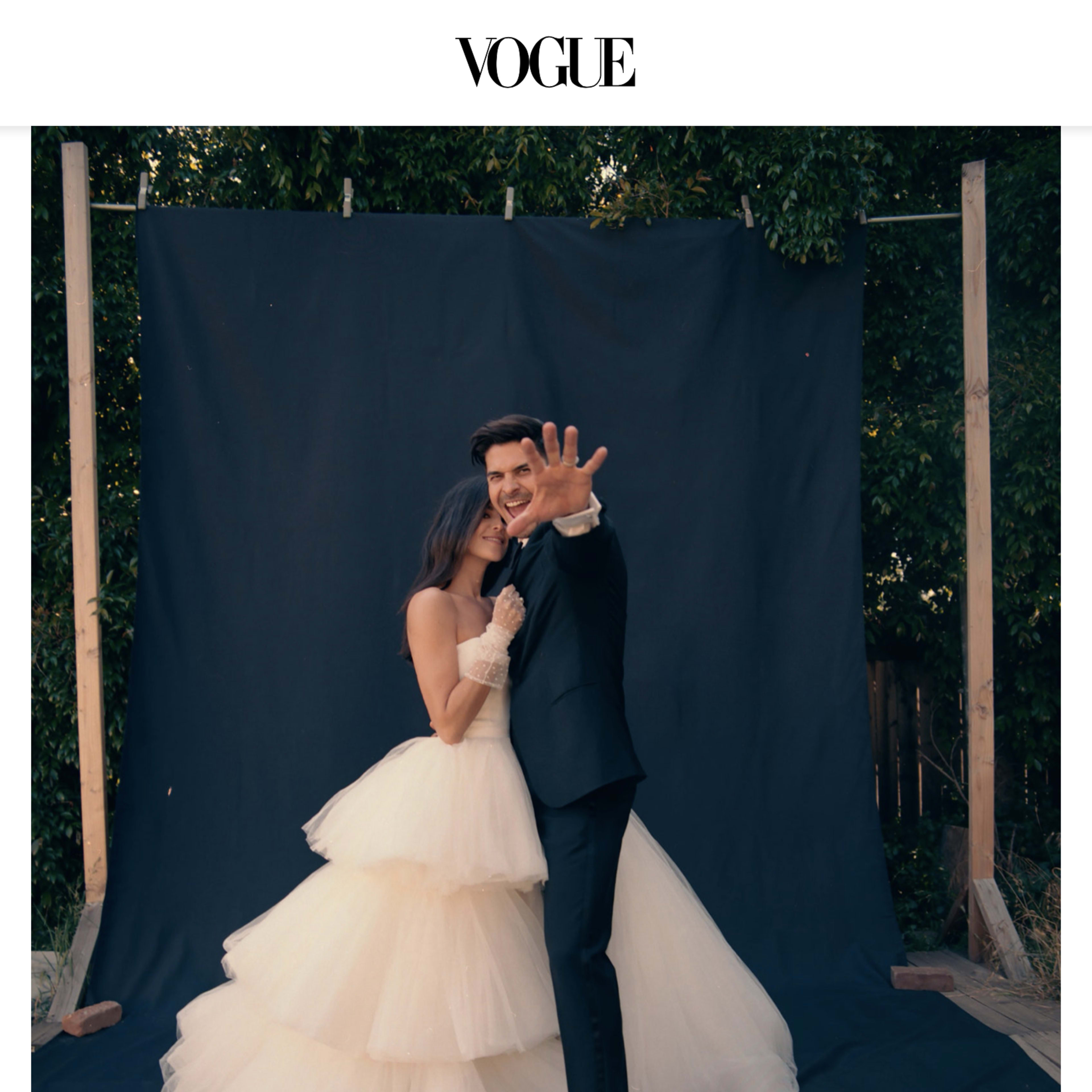 A couple poses in front of a dark backdrop, with the bride wearing a tiered wedding gown and the groom in a black suit, both smiling joyfully.