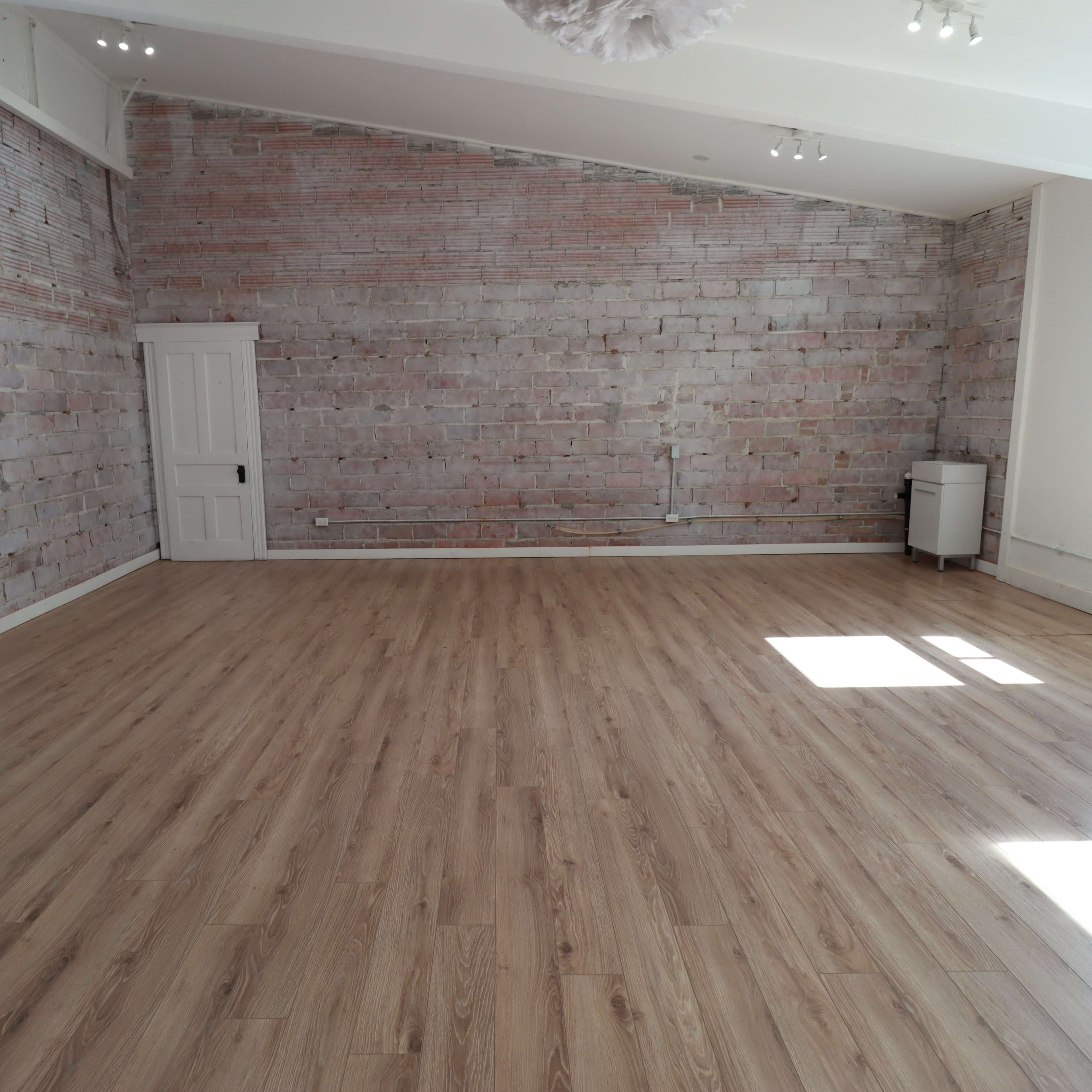 A spacious room with wooden flooring, exposed brick walls, a white door, and a small cabinet in the corner.