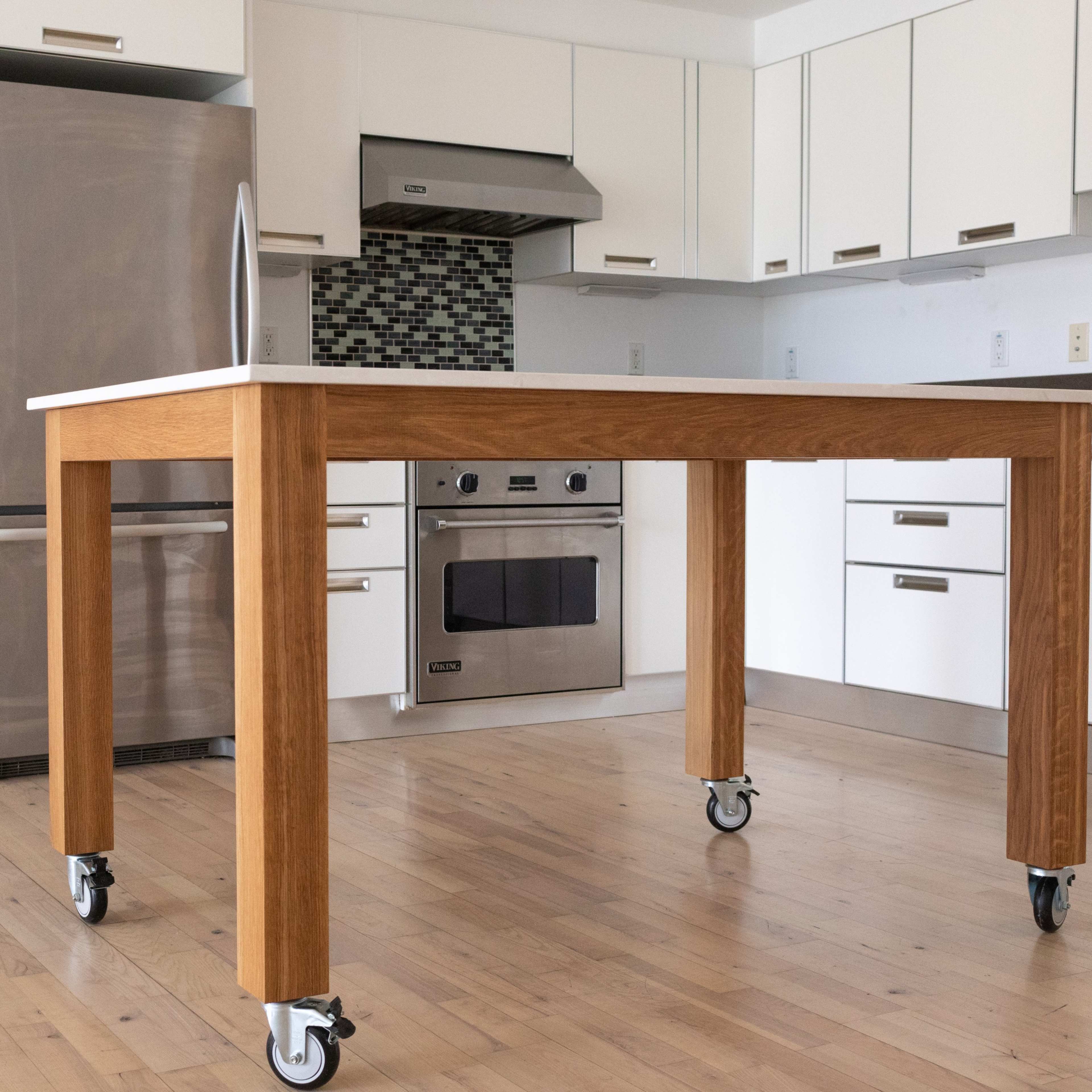 A wooden kitchen table on wheels positioned in a modern kitchen with stainless steel appliances and white cabinets.