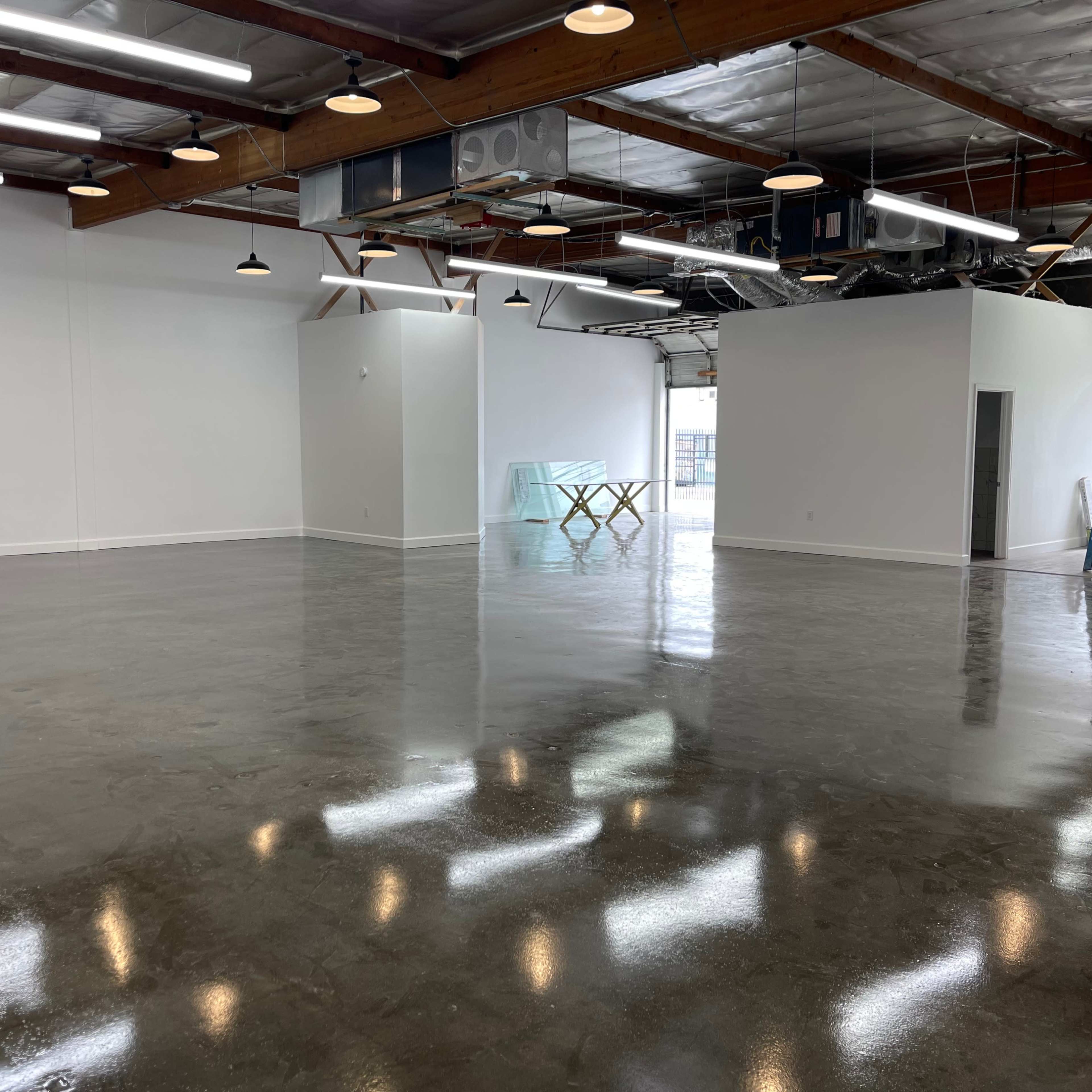 A spacious, empty industrial room with polished concrete floors and white walls, featuring wooden beams and several overhead lights.
