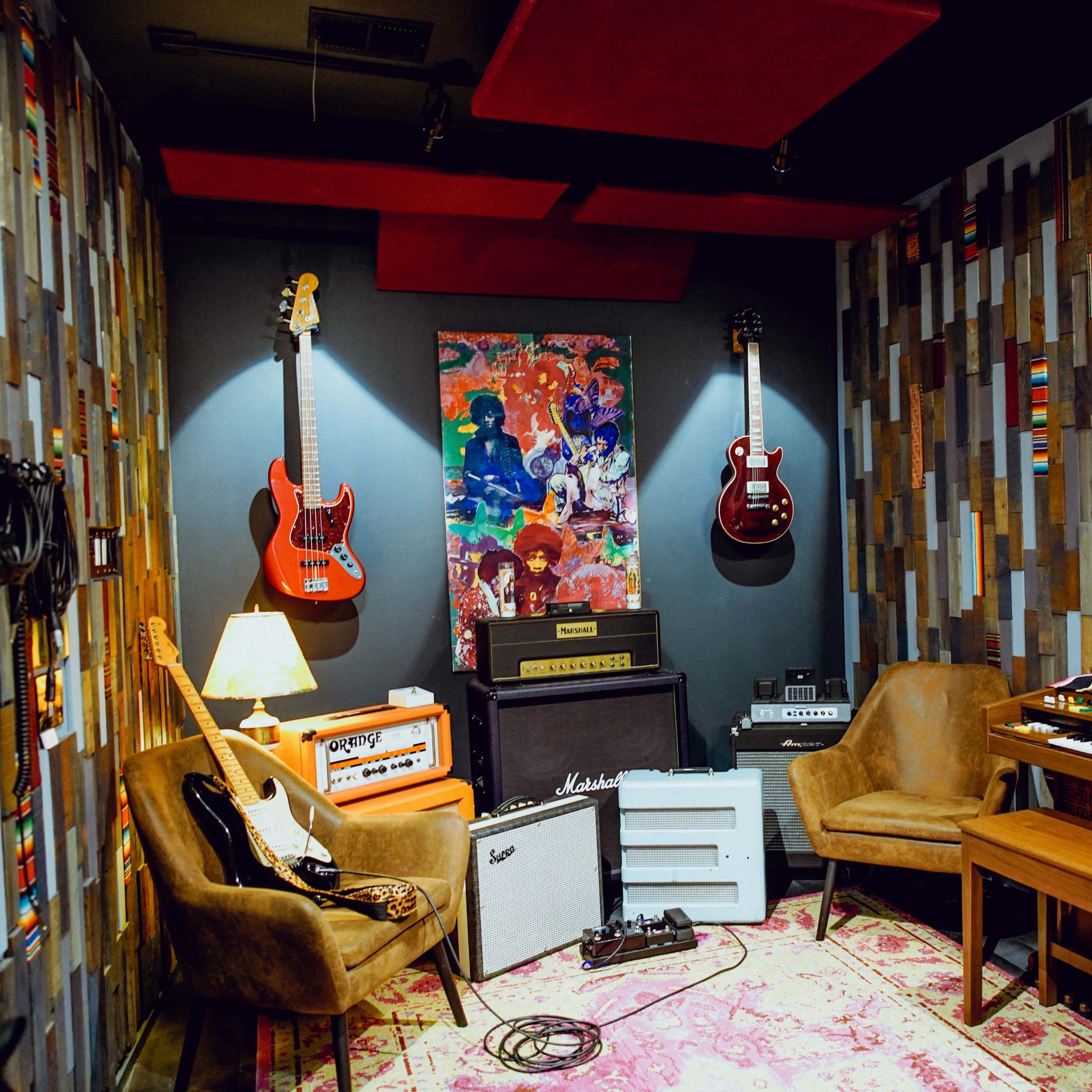 A music studio corner featuring two electric guitars on the wall, a vintage keyboard, amplifiers, and comfortable chairs, all set against a colorful patterned wall.