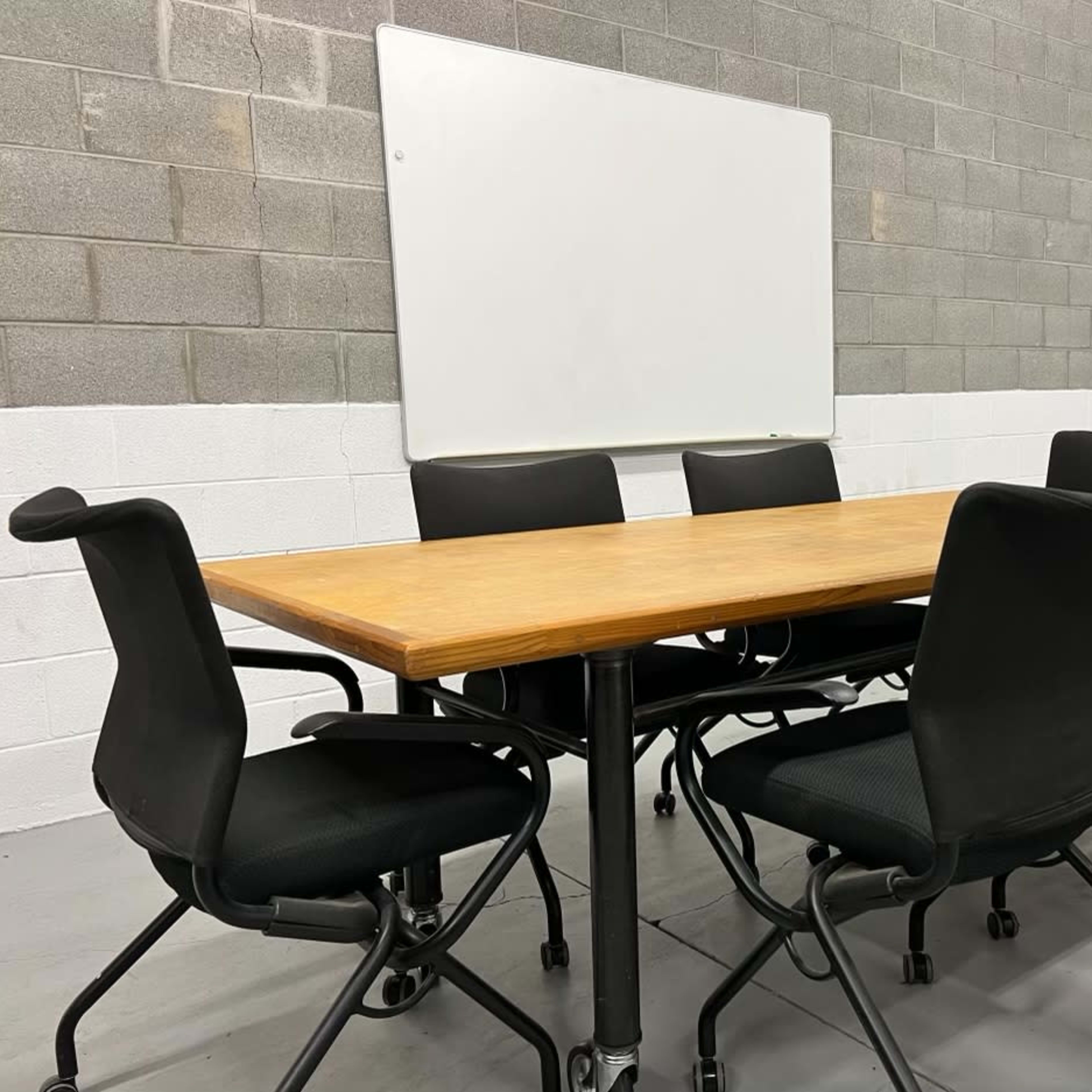 A rectangular wooden conference table is surrounded by six black rolling chairs in a room with gray block walls and a whiteboard mounted on the wall.