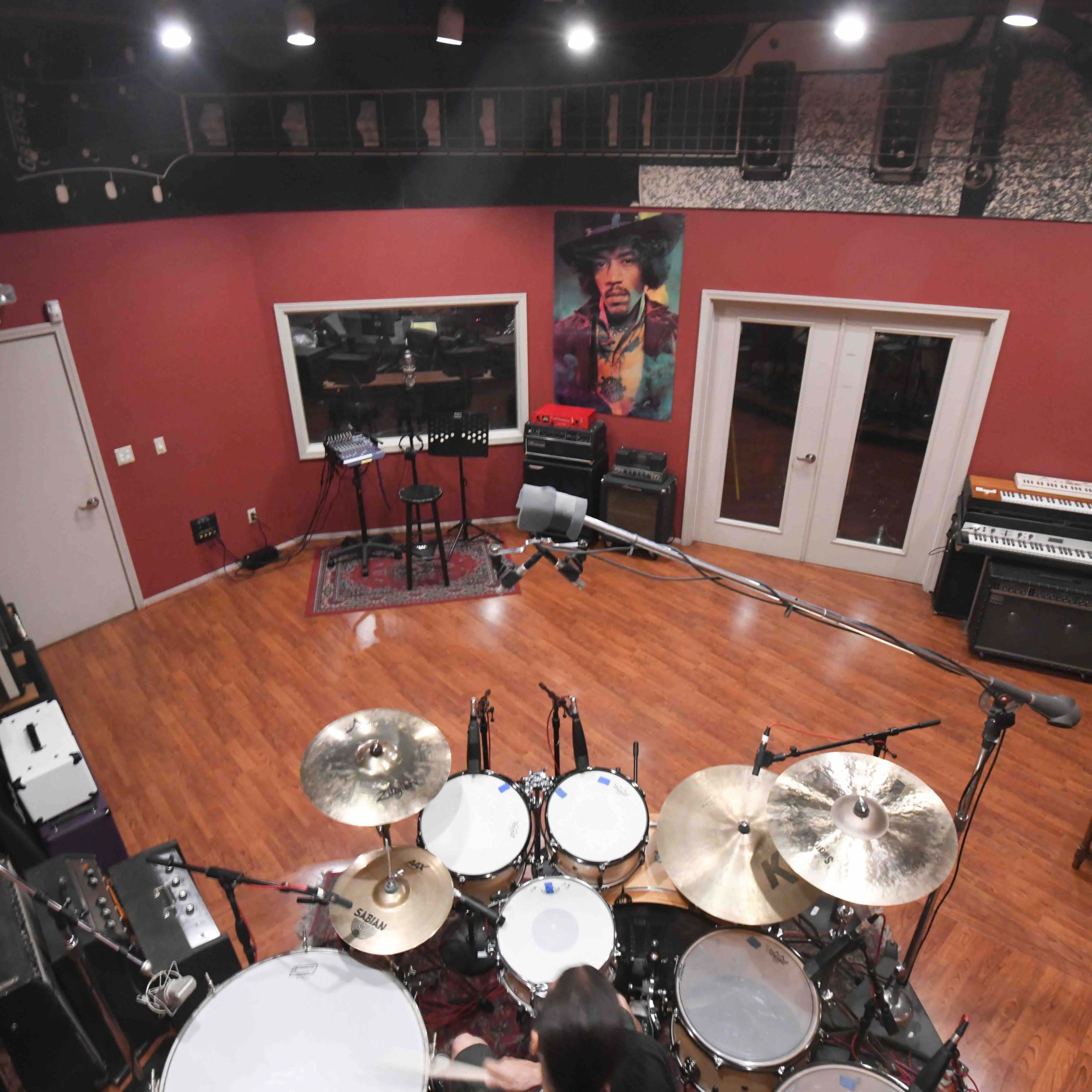 A well-equipped recording studio featuring a drum set in the foreground and various musical instruments along the walls, including a vintage keyboard and amplifiers.